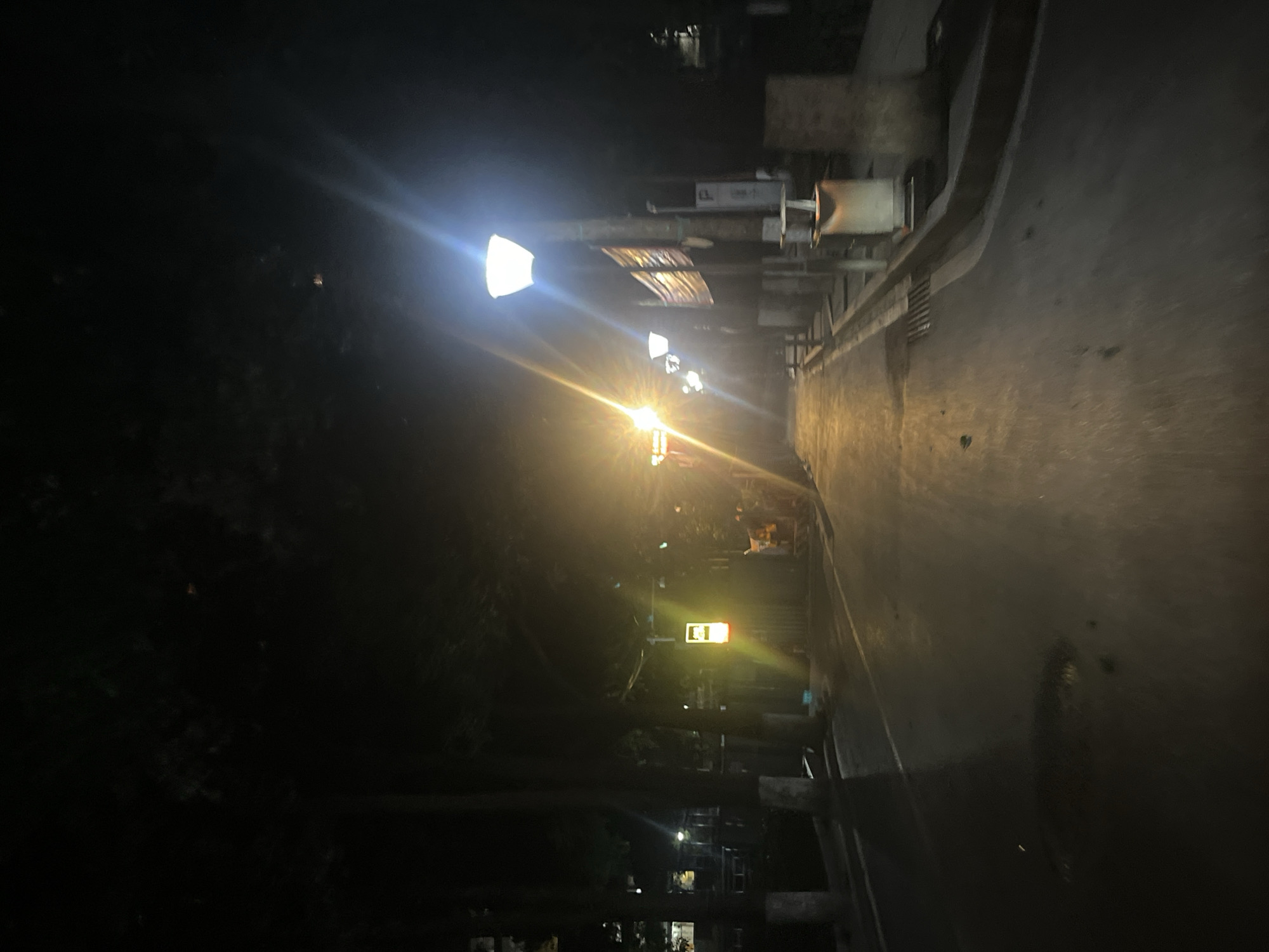 The image depicts a dimly lit street at night in Yuzhong District, China. A paved road occupies the foreground and midground, wet and reflective, suggesting recent rain or cleaning. On the right side of the road, a lamppost with a bright, white light illuminates the area. Further along the right side, there are several other streetlights and some planters or benches. A trash receptacle is visible near the curb. On the left side of the road, trees with dark foliage overhang the street, casting shadows. A few scattered lights from buildings can be seen in the distance. A yellow illuminated sign with a red and white element is visible on the left side of the road. The overall atmosphere is quiet and somewhat deserted, with the artificial lighting creating strong contrasts between light and shadow.