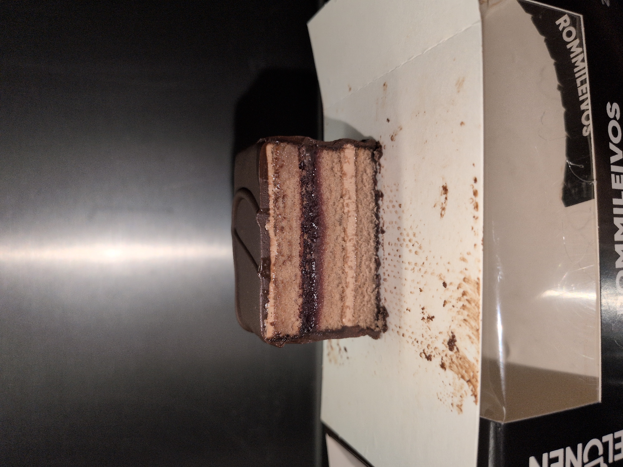A single rectangular slice of layered cake, identified as a "ROMMILEIVOS" (rum pastry) from the "ELONEN" brand, rests on a white, slightly soiled paper surface. The cake features a dark, glossy chocolate glaze on top, with a faint linear indentation. Its cross-section reveals three distinct layers of light brown cake sponge interspersed with two layers of light brown cream or mousse. A central, darker layer, consistent with fruit jam or a rich filling, is visible between the middle two cake layers. Dark chocolate crumbs and smears are scattered across the white surface, particularly in the foreground and to the left of the cake, indicating recent handling or consumption. The white paper surface is situated atop a dark cardboard box, where the product name and brand are visible. The background consists of a dark, reflective surface, possibly metal, displaying a prominent vertical strip of reflected light on the right side. No individuals are present in the image. The scene is consistent with a pastry being prepared for or consumed in an indoor setting within Vantaa, Finland, given the Finnish product labeling.