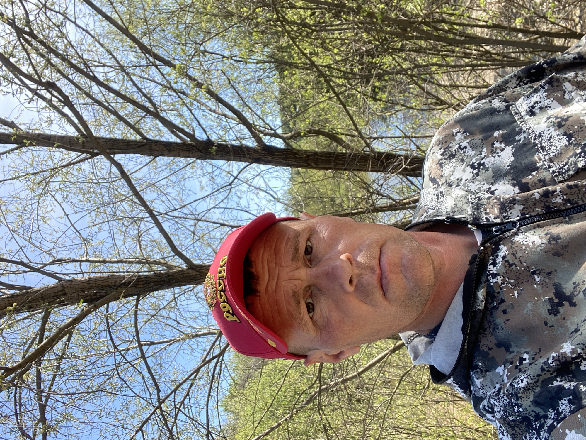 A man is photographed outdoors, likely during the daytime. He is wearing a red cap with gold-colored Cyrillic letters "РОССИЯ" (Russia) and a camouflage patterned jacket, partially visible. The background consists of bare tree branches and the sky. Based on the Cyrillic writing on the cap, the location is likely related to Russia. While the exact location within the city of Фоки, Russia cannot be determined solely from the image, the scene appears to be a natural environment, potentially a wooded area, park, or rural setting within or near the city.