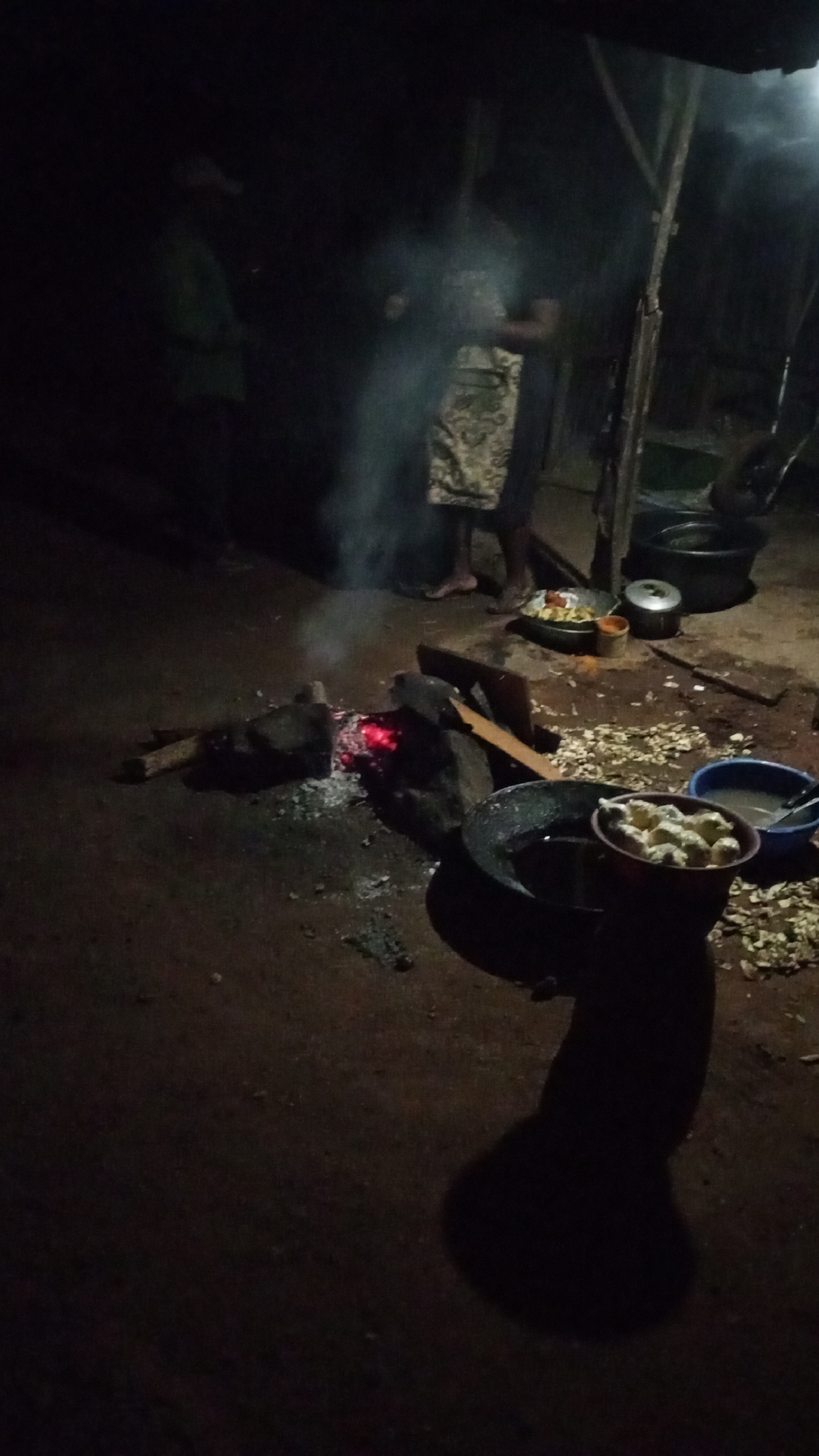 This media file captures an outdoor cooking scene at night in Sabon Tasha, Nigeria. The central focus is a small fire glowing with red embers on the unpaved ground, emitting wisps of smoke. This fire serves as a heat source for cooking.
To the right of the fire, various cooking items are arranged on the ground. A large, dark pan, possibly containing oil for frying, is positioned near the embers. Adjacent to it, a red bowl holds a quantity of light-colored, irregularly shaped food items, possibly dough balls or fritters, ready for cooking. A blue basin containing liquid and other utensils is also visible. Further back, near a standing figure, additional food preparation elements include a tray with peeled food items (like yams or potatoes) and a lidded pot. Scattered white scraps on the ground suggest ongoing food preparation.
Two figures are present in the dim light. On the left, a person wearing a light-colored top and a hat stands partially obscured by shadows. On the right, closer to the cooking area, another individual stands, wearing what appears to be an apron or patterned fabric around their waist, with their bare feet visible. Both figures appear to be engaged in or observing the cooking process. The background features simple, makeshift wooden structures. The scene conveys a communal or household activity taking place under the cover of darkness, with the fire providing the primary illumination. No text is visible in the image.