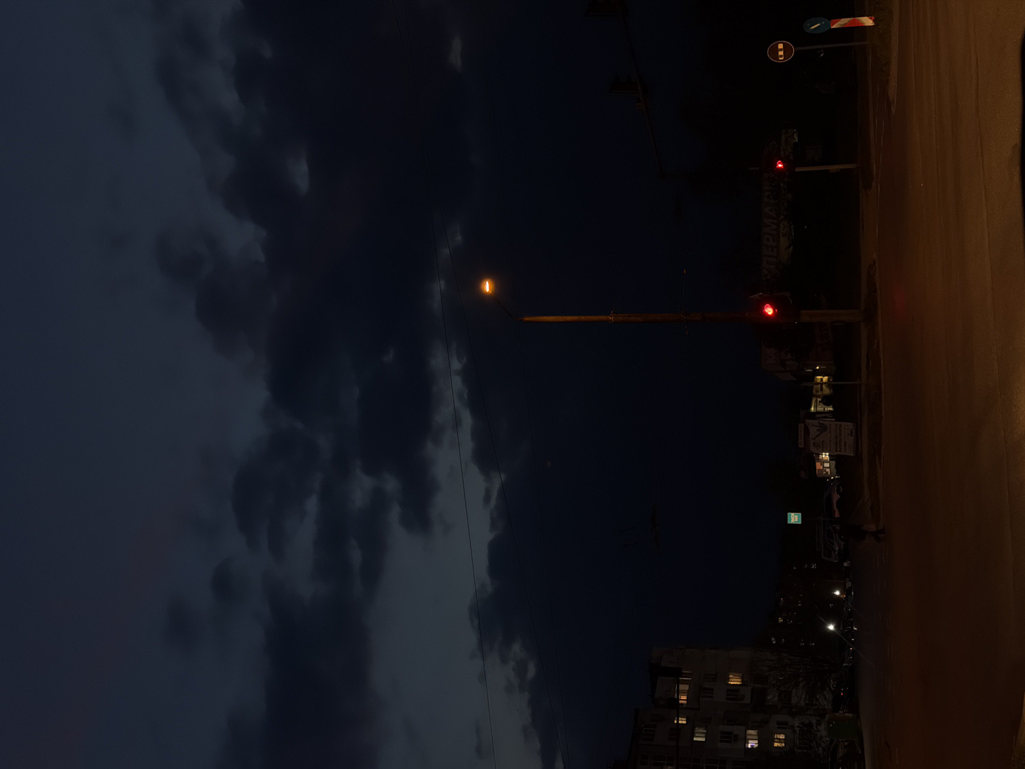 The image displays an overcast night scene within an urban environment, consistent with Varna, Bulgaria. The sky, occupying the upper and left portions of the frame, is dark blue-gray and filled with heavy, dark clouds.
A single, illuminated yellow-orange streetlight on a pole dominates the mid-left, casting light downwards. On the right side, two red traffic lights are visible, indicating a stop signal for vehicles; one is positioned higher on a pole, and another lower. Several dark power lines traverse the sky.
The right side of the image features a dark, broad road or pavement surface, running vertically and subtly reflecting the artificial lights. Near the top right, two road signs are present: a circular "No Entry" sign (blue with a white horizontal bar) and an indistinct red and white rectangular sign.
In the lower and right sections, buildings with some illuminated windows are visible, suggesting activity within. Faint Cyrillic text, appearing to be "СУПЕРМАРКЕТ" (SUPERMARKET), can be seen on a sign or building facade behind the upper traffic light, providing a specific location cue.
The scene is static, devoid of clearly discernible people or moving vehicles. The overall atmosphere is that of a quiet, dark city night under heavy cloud cover, illuminated by artificial light sources.