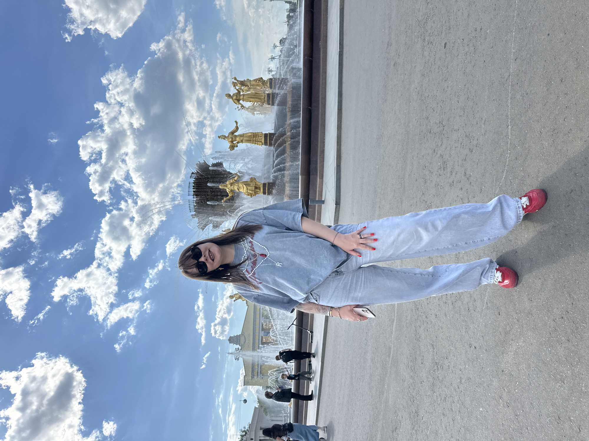 A young woman stands centrally in a public outdoor space on a bright, sunny day in what appears to be Moscow, Russia. She is facing slightly towards the viewer, with a relaxed posture and a subtle smile. She has long brown hair with bangs, wears dark, round sunglasses, a light gray oversized t-shirt, light blue loose-fitting jeans, and striking red sneakers with white laces. Her left hand, adorned with red nail polish and several rings, holds a white smartphone. A distinctive white and red beaded necklace with a silver 'R' pendant hangs around her neck.
Behind the woman, a large and ornate golden fountain dominates the scene. The fountain features multiple tiers with numerous golden statues of figures, likely allegorical, spraying water. The water glistens under the bright sun. Beyond the fountain, in the distance, a grand building with columns is visible, further hinting at a significant public landmark. Several other people, mostly seen as dark silhouettes, are scattered around the fountain area, some observing it or walking by, with one pushing a stroller. The ground is paved, appearing as a light gray asphalt or concrete surface.
The sky is a vibrant blue, filled with scattered white, fluffy cumulus clouds, indicating fair weather. The strong light suggests it is midday or early afternoon. The specific design of the fountain, known as the "Friendship of Nations" fountain, along with the architectural style of the distant building, points to VDNKh (Exhibition of Achievements of National Economy) in Moscow as the location. The overall mood conveyed is one of leisure and enjoyment of a beautiful public park setting.