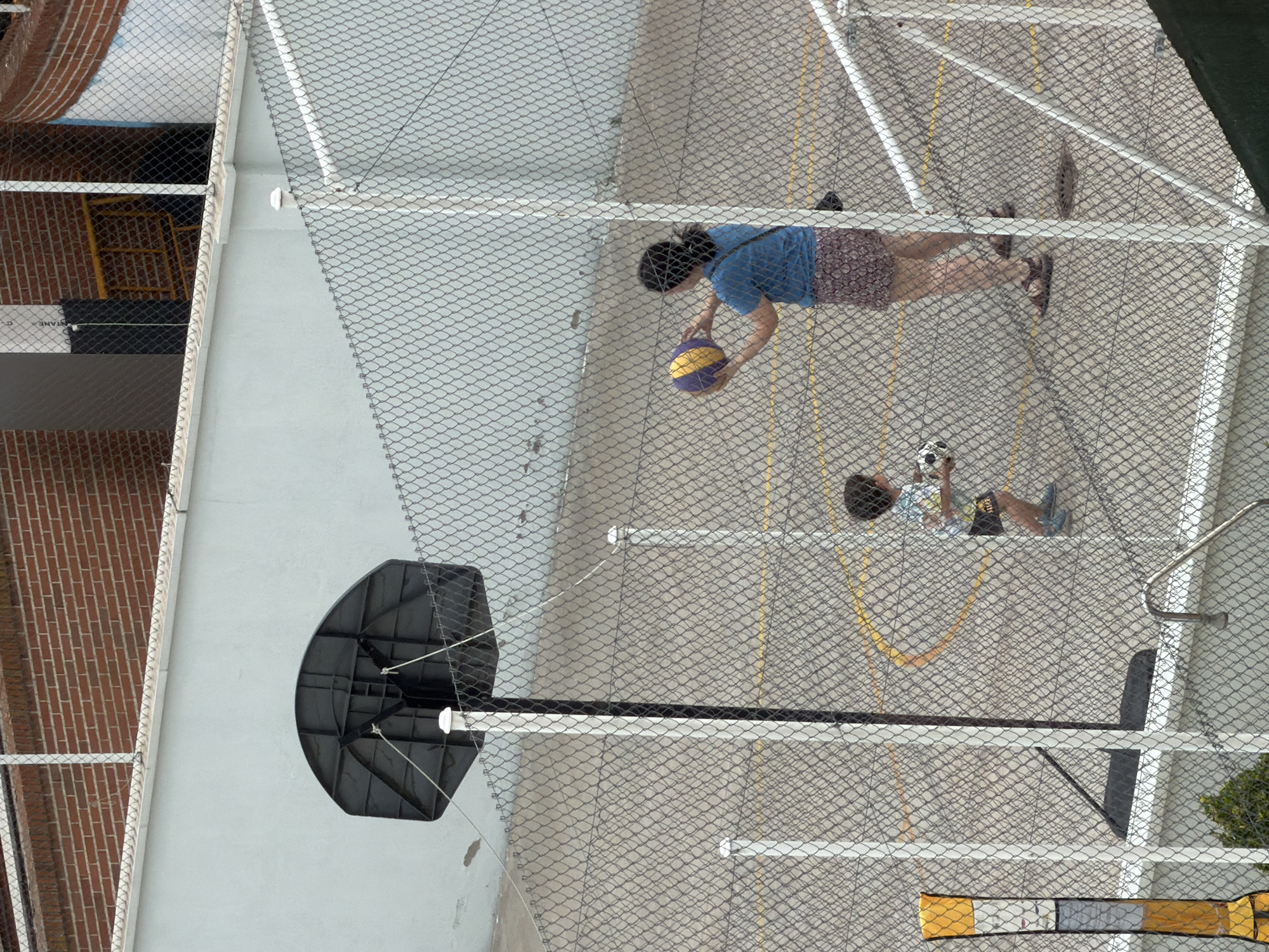 An aerial, medium shot captures a fenced outdoor basketball court with two people on it. To the right, a woman with dark hair, wearing a blue t-shirt and a patterned skirt, holds a yellow and purple ball. To her left, a young child in a t-shirt and shorts stands with a ball. A basketball hoop with a black backboard is visible on the left side of the frame. The court is surfaced with asphalt and marked with yellow lines. A chain-link fence encloses the court, with a brick building and a lighter colored wall in the background. The scene appears to be an urban recreational area.
