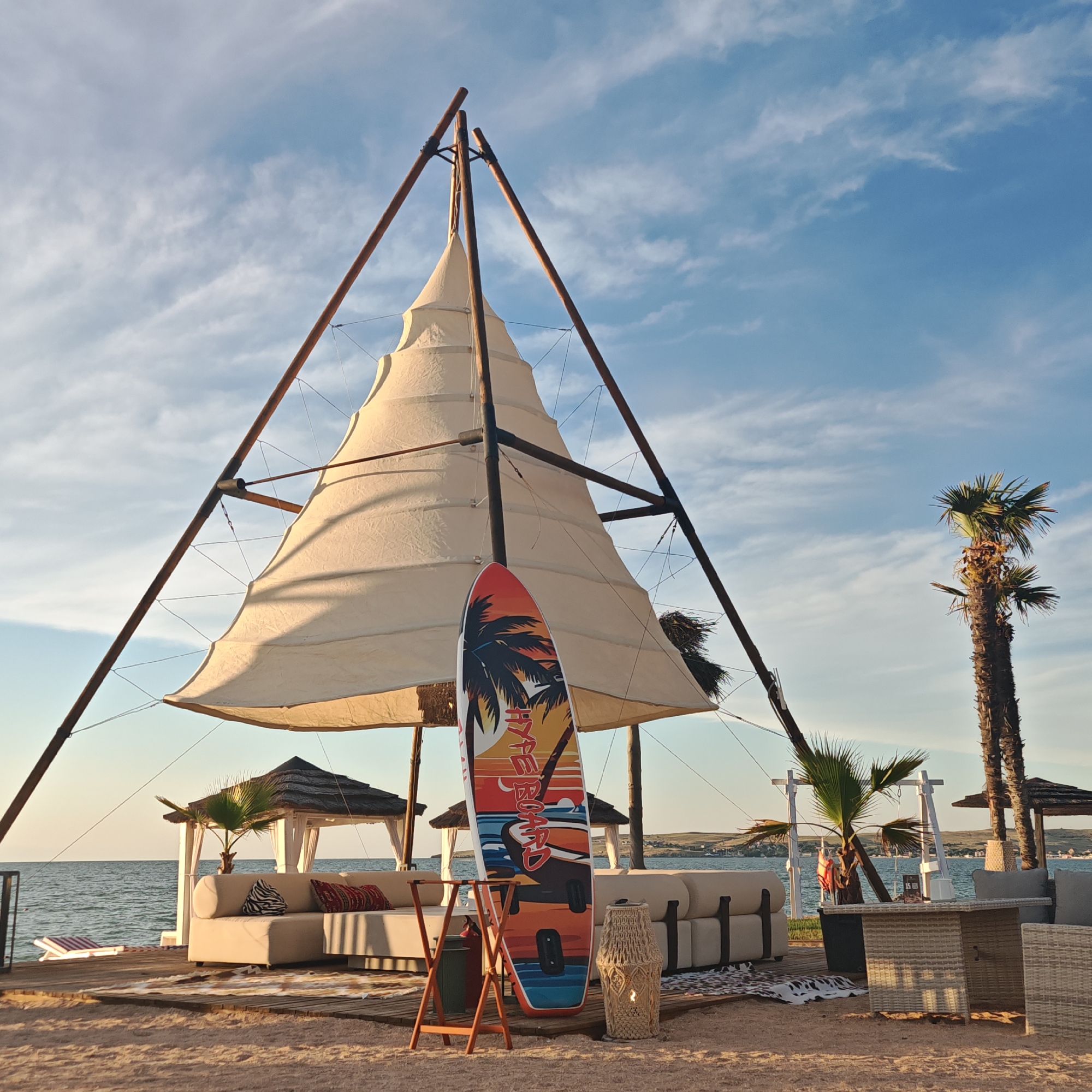 The image depicts a beach scene at Мысовское сельское поселение, Ukraine. A large, sail-like structure made of off-white fabric is supported by wooden poles. In front of this structure, a surfboard with a tropical sunset design and the words "HYPE BOARD" is propped up on a wooden stand. To the right of the surfboard, a wicker lamp with a lit candle sits on the sand. Behind these items, a lounge area is set up on a wooden platform, featuring white sectional sofas with decorative pillows and a wicker coffee table. Two thatched-roof gazebos are visible behind the sofas. The scene is on a sandy beach with the sea in the background. Two palm trees are present, one taller and more prominent on the right. The sky is blue with scattered white clouds. The sun appears to be setting or rising, casting a warm light on the scene. There are no people visible in the image.