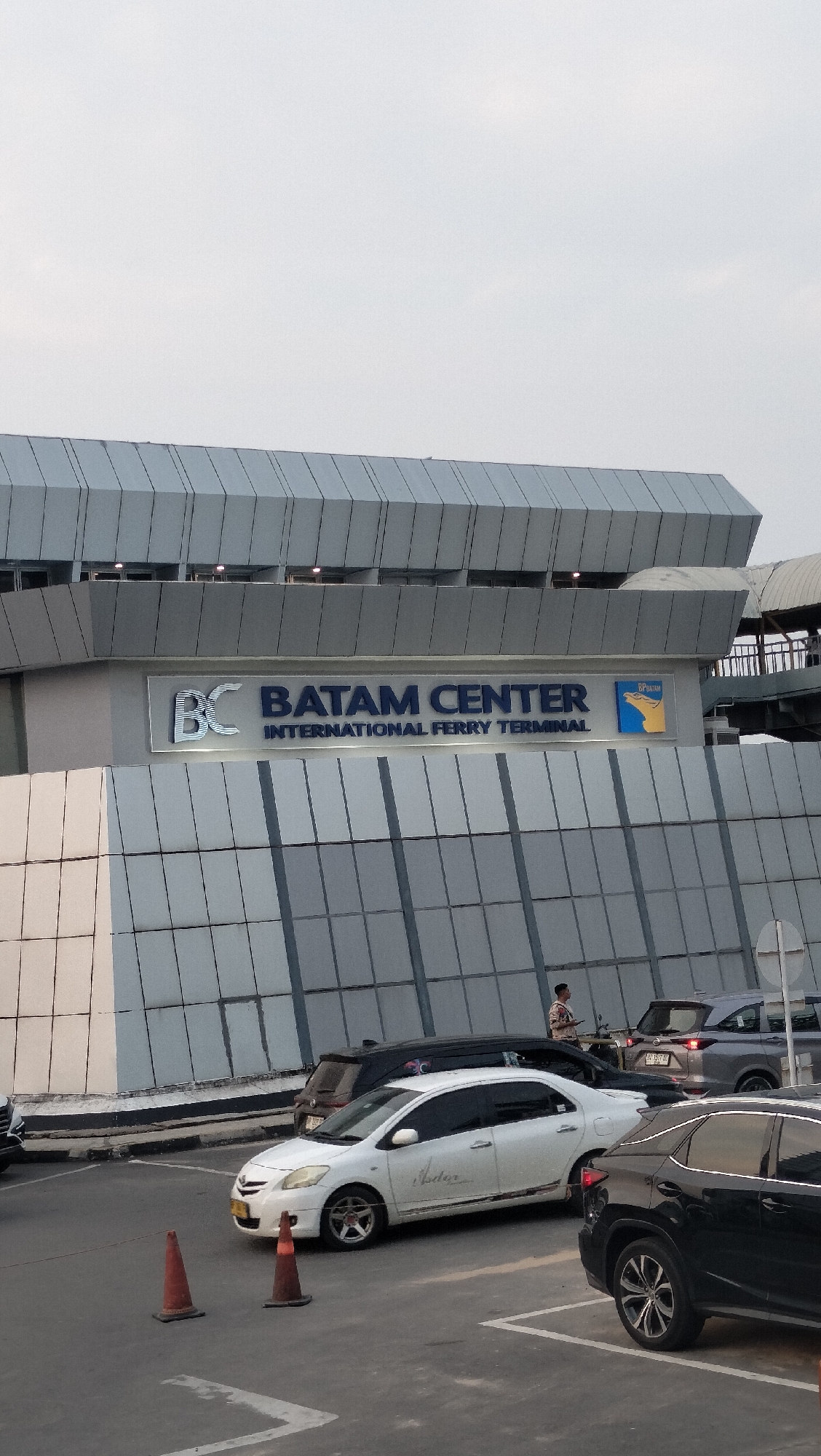 The image shows the exterior of the Batam Center International Ferry Terminal in Batam, Indonesia. The building has a modern architectural design with a gray, segmented facade. Prominently displayed on the building is the text "BC BATAM CENTER INTERNATIONAL FERRY TERMINAL" in large, blue, illuminated letters. To the right of the text is a logo featuring a stylized depiction of a bird in flight, colored yellow and blue, with the text "BP BATAM" above it.
In the foreground, a paved area, likely a parking lot or drop-off zone, is visible. Several cars are present, including a white sedan, a black SUV, and a dark-colored minivan. Two orange traffic cones are placed on the asphalt. One person, appearing to be a man wearing a patterned shirt, is standing near the vehicles, possibly a passenger or employee. The sky is overcast, suggesting a daytime scene. The overall environment appears to be an active transportation hub.