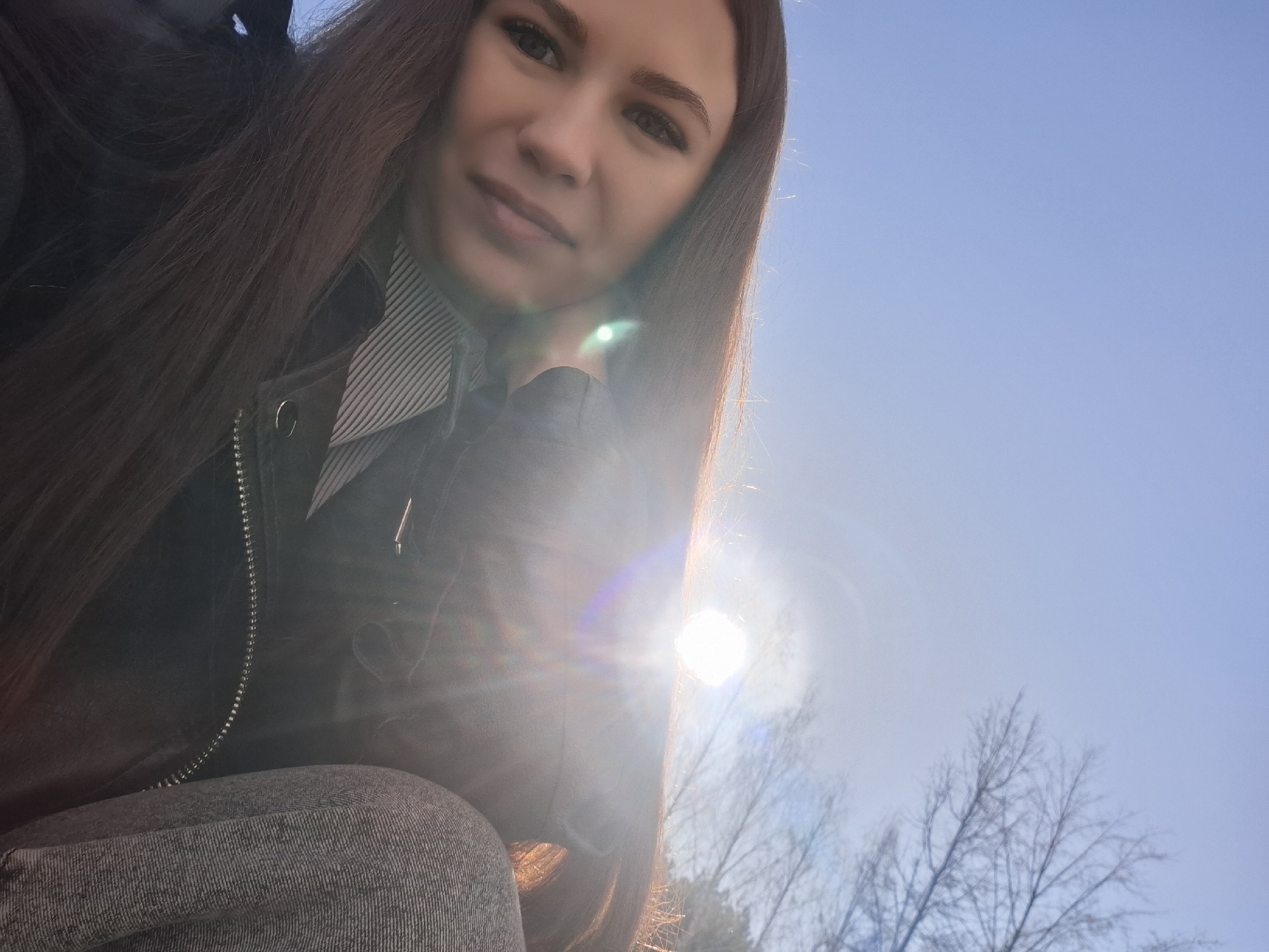 A selfie of a woman with long brown hair, wearing a black jacket over a striped shirt, taken outdoors on a sunny day. The background features a bright blue sky and the silhouette of leafless trees. The location appears to be Chaikovsky, Russia. The bright sun creates a lens flare effect.
