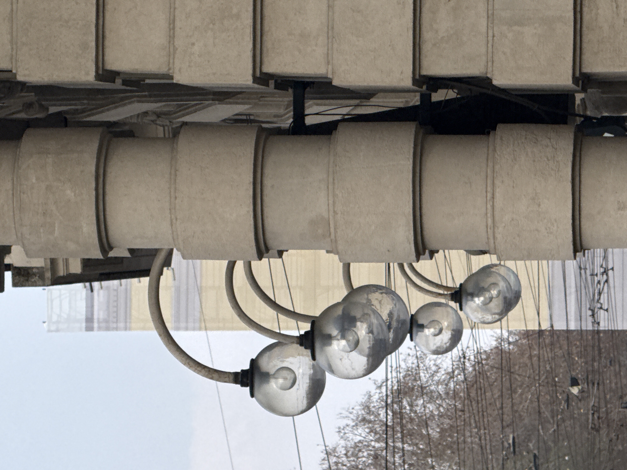 The image presents a detailed view of an urban environment in Milan, Italy, focusing on architectural features and street infrastructure. The prominent subject is a light-colored building facade, likely composed of stone or concrete. This facade features a vertical stack of cylindrical sections on the left, resembling a column, and an adjacent section of rectangular, ashlar-like blocks on the right, separated by a deep, dark vertical recess. Four streetlights are horizontally mounted at regular intervals on the cylindrical facade section. Each streetlight consists of a curved, dark-colored arm supporting a spherical, textured glass globe containing an incandescent bulb. The globes reflect ambient light and distorted elements of the surroundings. To the left, a leafless deciduous tree partially fills the mid-ground. Numerous dark, thin overhead wires, consistent with public electric transportation infrastructure, traverse the upper-mid section of the frame. In the background, under an overcast sky, a taller building partially clad in white scaffolding netting is discernible. No people are visible, and no dynamic actions or direct interactions are depicted.