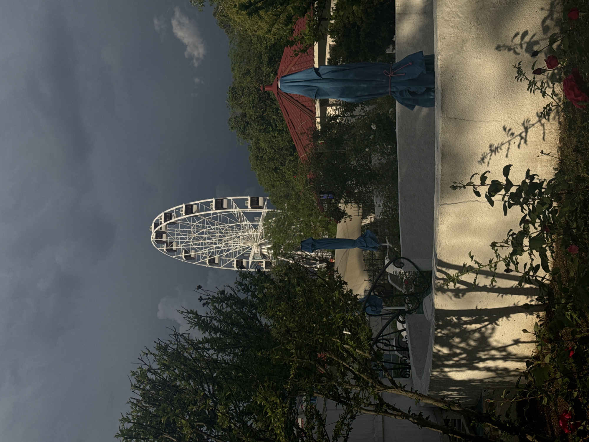 The image shows a Ferris wheel against a cloudy sky, partially obscured by trees. In the foreground, a white wall and patio umbrella are visible, along with some plants and decorative metalwork. Based on the presence of the Ferris wheel, the location is likely Yevpatoriya, Ukraine.