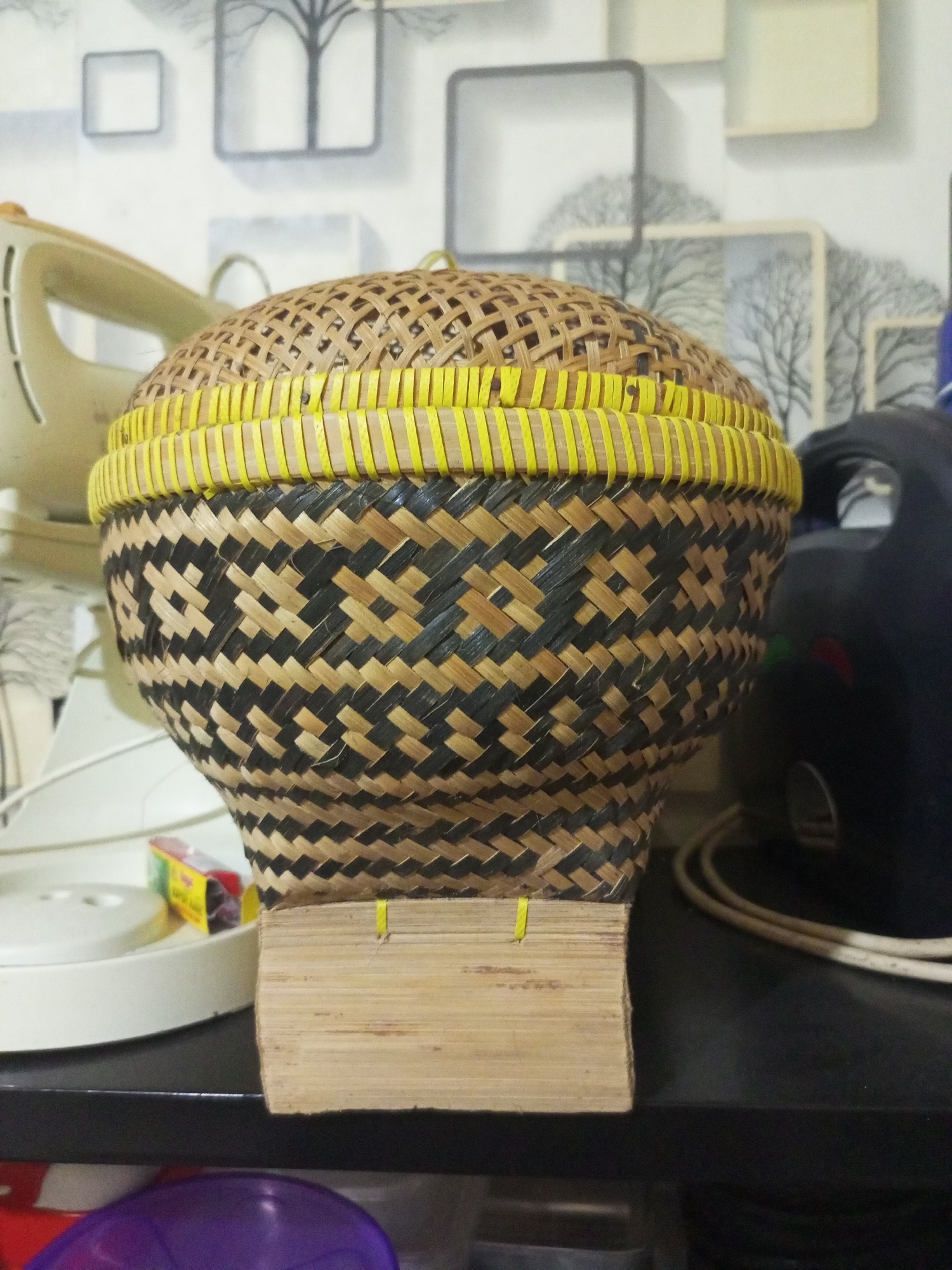 A tightly woven, conical basket with a lid made of bamboo and yellow accents is centrally placed on a dark shelf. The basket features a black and natural woven pattern around its body. Attached to the front of the basket's base is a small, rectangular piece of light-colored bamboo, also secured with yellow string. To the left of the basket, a portion of a cream-colored electrical appliance, possibly a fan or heater, is visible, with its base resting on the same shelf. Further to the left and below, a white circular object and a small yellow and red box are partially seen. To the right of the basket, a large, dark object, potentially another appliance with a handle, and some coiled white electrical cords are present on the shelf. The background consists of a light-colored wall with a repeating wallpaper pattern of stylized trees and geometric shapes. The overall scene appears to be an indoor setting, likely a storage area or a room within a home or shop in Cidahu, Indonesia. There are no people or animals visible in the image.
