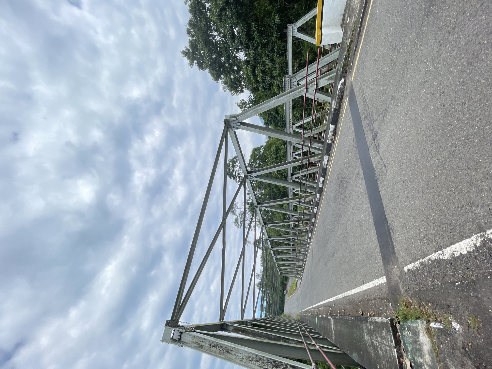 The image shows a view from the side of a bridge with a metal truss structure. The bridge has an asphalt road with white lines. On the right side of the road, there are some yellow and white concrete barriers. Lush green trees are visible beyond the bridge. The sky is cloudy. The scene appears to be in a rural or semi-rural area.
