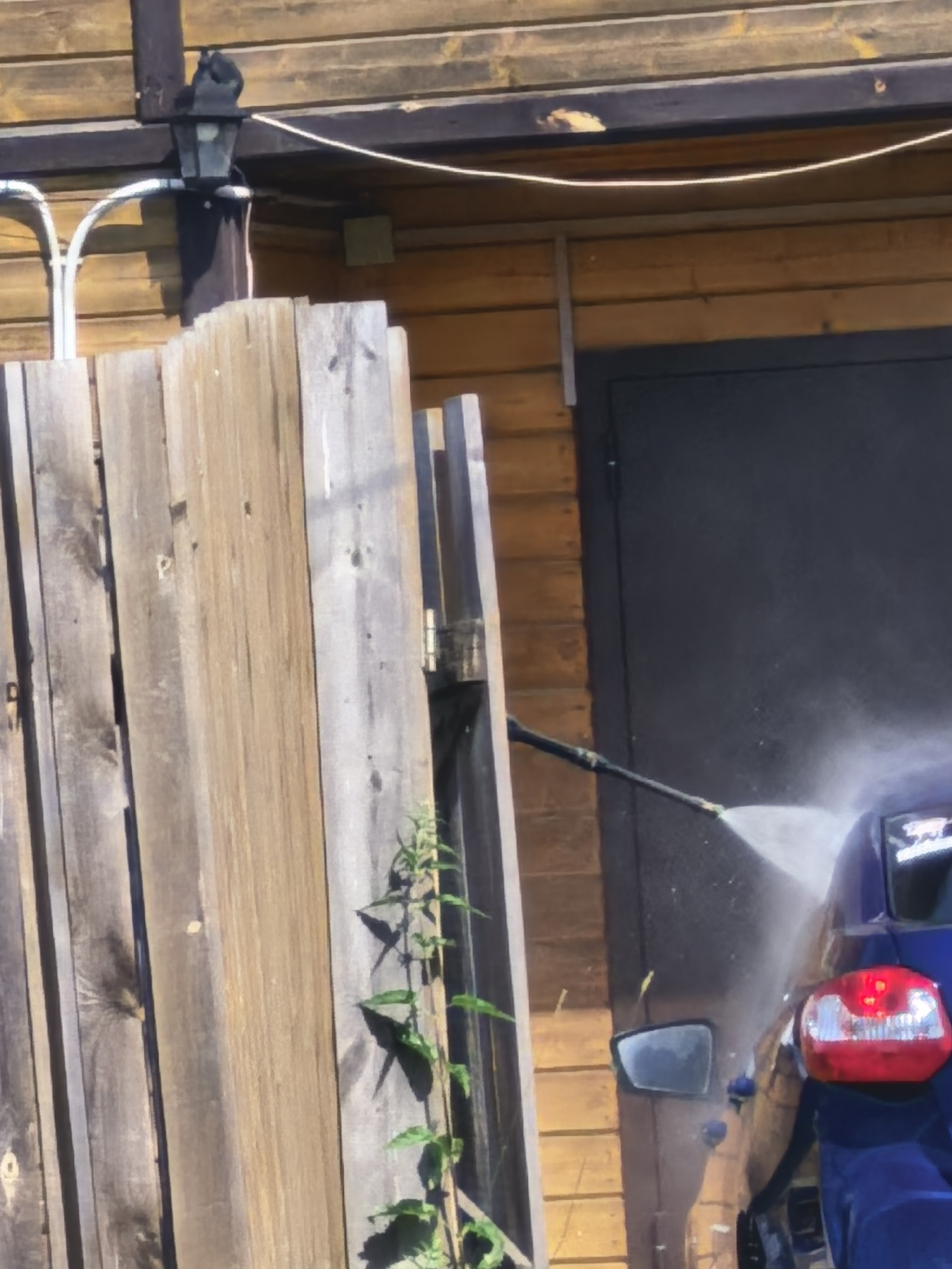 A pressure washer is being used to clean a blue car parked next to a wooden fence and a wooden building. The car's taillight is visible, and a spray of water is obscuring part of the vehicle and the building. A black side mirror is also visible on the car. The building appears to be a garage or shed with a dark colored door. The fence is made of weathered wooden planks.