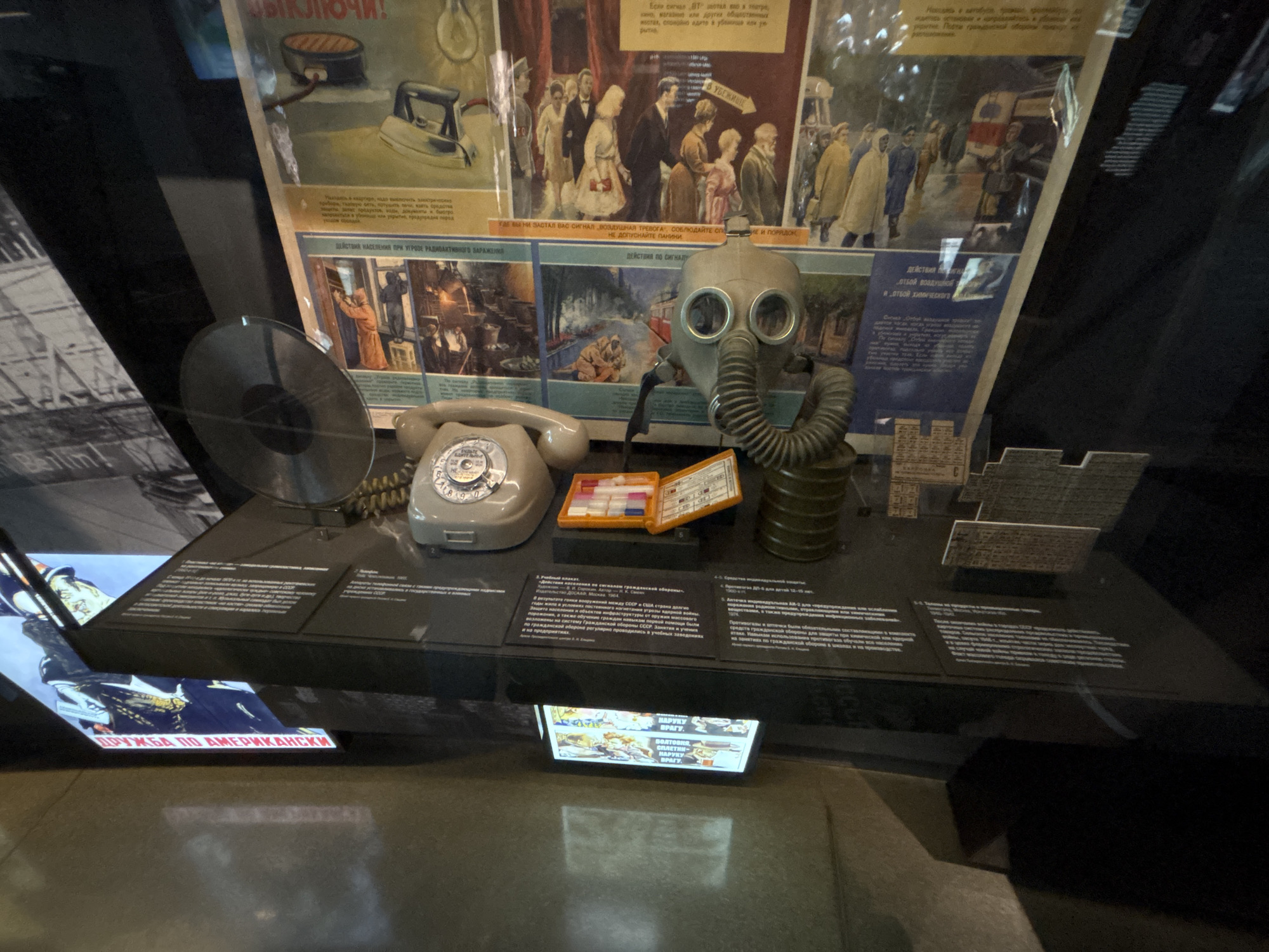 This image captures a museum display case showcasing Soviet-era civil defense and domestic artifacts, located in Yekaterinburg, Russia.
The upper background features three faded Soviet propaganda posters. The leftmost depicts an electric kettle and an iron with the warning "ВЫКЛЮЧИ!" (Turn off!). The central poster shows formally dressed individuals walking, with text partially obscured but including "В УБЕЖИЩЕ!" (To the shelter!). The rightmost poster displays people in a city setting.
The lower background presents three additional informational posters. The left poster, titled "ДЕЙСТВИЯ НАСЕЛЕНИЯ ПРИ УГРОЗЕ РАДИОАКТИВНОГО ЗАРАЖЕНИЯ" (Actions of the Population During the Threat of Radioactive Contamination), illustrates individuals securing windows. The central poster, "ДЕЙСТВИЯ ПО СИГНАЛУ" (Actions by Signal), depicts people taking cover in a natural environment. The rightmost poster, "ОТБОЙ ВОЗДУШНОЙ" (All Clear Air), shows people in winter clothing.
Within the display case, from left to right, are a black vinyl record, a gray rotary-dial telephone with a coiled cord, an open orange plastic first-aid kit containing various small pill containers, a gray Soviet GP-5 gas mask with circular eye lenses and a corrugated breathing tube connected to a green cylindrical filter canister, and several small, rectangular metal or plastic elements. Dark informational panels with white Russian text are positioned in front of the artifacts.
Below the main display, a glowing digital screen displays additional images and text, including phrases such as "ДРУЖБА ПО АМЕРИКАНСКИ" (Friendship, American Style) and "ВРАГУ!" (To the enemy!), indicating themes of Cold War propaganda. The entire exhibit is enclosed within a glass display case, exhibiting reflections from the museum environment and overhead lighting.