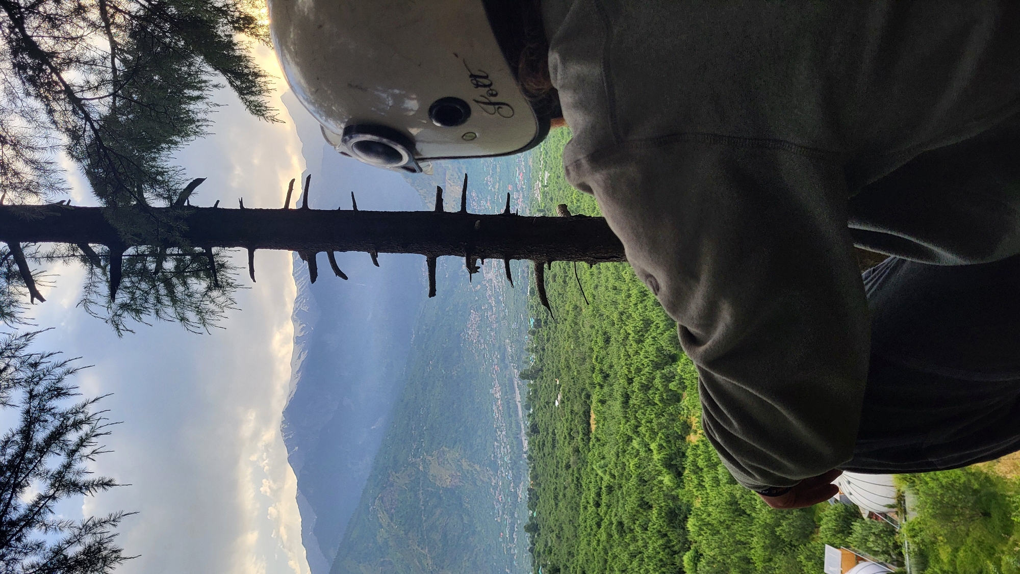 The image shows a first-person perspective of a person wearing a helmet and seated, likely on a platform, overlooking a landscape in Nashala, India. The foreground includes a tree trunk with sparse branches, situated between the viewer and the expansive view. The background features a valley with lush green vegetation, and possibly residential areas, leading up to a mountain range visible under a sky with scattered clouds. The light suggests either early morning or late afternoon. The individual is wearing a hooded sweatshirt. The helmet is white with lettering "Jett".