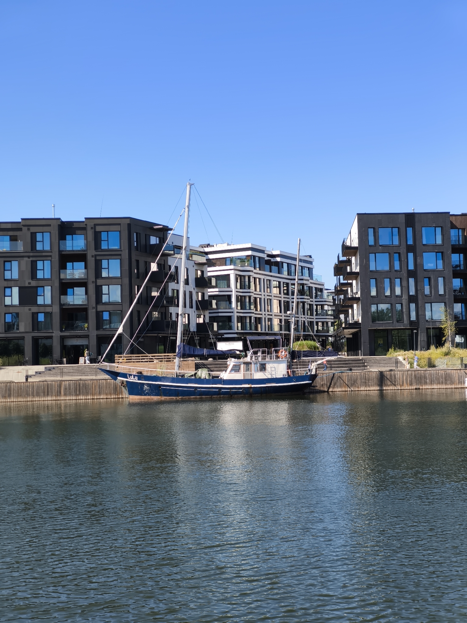 The image depicts a blue sailboat docked in a harbor with modern, multi-story buildings in the background. The buildings are a mix of dark gray and white facades with numerous glass windows, suggesting an urban waterfront area. The sky is clear and blue. The sailboat has a white mast and rigging, and its hull is painted dark blue with "Lulu" written in white lettering on its side. The harbor water is calm, reflecting the sky and buildings. Several people can be seen walking along the waterfront promenade in front of the buildings. The scene appears to be in Tallinn, Estonia, specifically the Noblessner Quarter, a developing waterfront area known for its residential and commercial buildings.