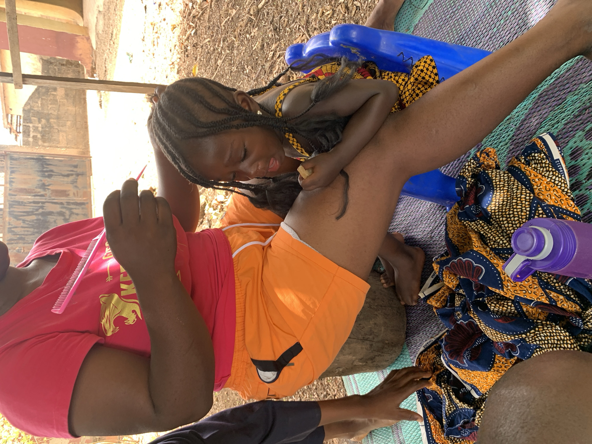 The image, possibly taken in the city of Akwanga, Nigeria, shows an outdoor scene. An adult wears a pink t-shirt with a gold design and orange shorts. The adult sits on a log and holds a pink comb in their right hand. A young child with braided hair lies across the adult's lap. The child wears a yellow and black patterned top and holds something small in their hand. A blue plastic object is near the child, possibly a small chair or toy. A colorful patterned fabric and a purple water bottle are on a woven mat under the adult and child. The environment appears to be shaded, with dappled sunlight filtering through foliage. A building is visible in the background.