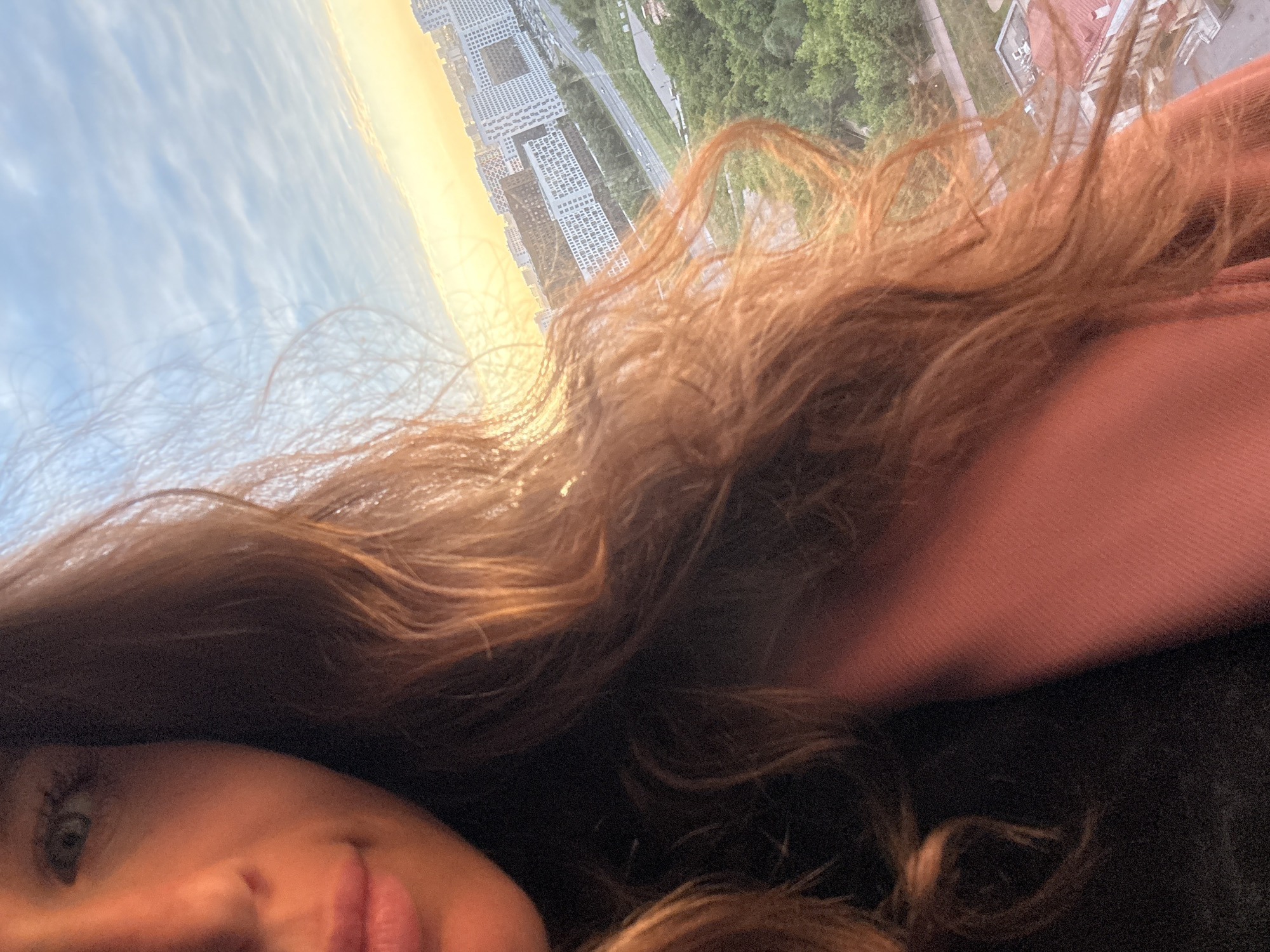 The image is a selfie taken by a woman in Dolgoprudny, Russia, with a cityscape in the background. The woman, with light skin and long, wavy auburn hair, is positioned on the left side of the frame, with her face and upper torso visible. She is wearing a dark top and a pinkish jacket. Her gaze is directed towards the camera, and she has a subtle smile.
The background depicts a residential area of Dolgoprudny, characterized by numerous multi-story buildings, some with modern architectural designs and white facades. A road with vehicles is visible, running through the urban landscape. Lush green trees and vegetation are interspersed between the buildings, suggesting a suburban or park-like setting within the city. The sky is partly cloudy with hints of sunset hues, casting a warm light on the scene. The overall impression is that of a casual, everyday moment captured in an urban environment.