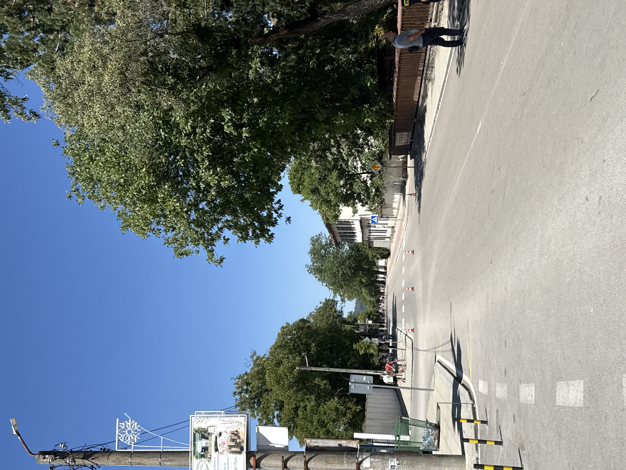 The image depicts a street scene in the city of Oreshak, Bulgaria, on a clear, sunny day. The perspective is from the street, looking down a road lined with trees and buildings.
On the left side of the street, a concrete utility pole with electrical wires and a street lamp is visible. Attached to the pole is a white, snowflake-shaped decoration and a framed sign with an image and text. Further down the street on the left, a green trash receptacle is filled with plastic bottles. A group of individuals, appearing to be tourists, are walking on the sidewalk. Several orange traffic cones are placed along the road.
The road itself is paved and appears to be a two-lane street. White dashed lines mark the lanes, and a pedestrian crossing is visible.
On the right side of the street, a white building with dark shutters is situated behind a low wall. Further along, a wooden fence with a decorative railing runs along the sidewalk. Two individuals, one wearing a grey t-shirt and dark pants, and another partially visible person in dark clothing, are standing near the fence. A large tree with dense green foliage dominates the right side of the frame, casting shadows on the road.
The sky is a clear, bright blue. The overall impression is that of a quiet, residential or commercial street on a warm day.