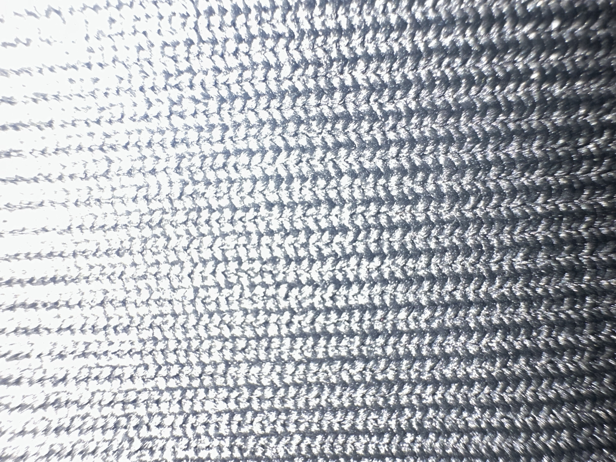 The image displays a close-up view of a textured, knitted fabric, likely a textile material. The fabric exhibits a repeating pattern of interlocking stitches, creating a chevron-like or herringbone design. The color is predominantly silver or metallic gray, with variations in light and shadow suggesting a subtle sheen or reflective quality of the material. The texture is finely detailed, with individual threads or loops visible.
There is no discernible location within the city of Letterkenny, Ireland, depicted in this image. The image focuses solely on the fabric's texture and pattern, lacking any contextual elements that would identify a specific place. No people, subjects, objects, actions, or interactions are present in the scene. The environment is abstract, reduced to the surface of the fabric itself.