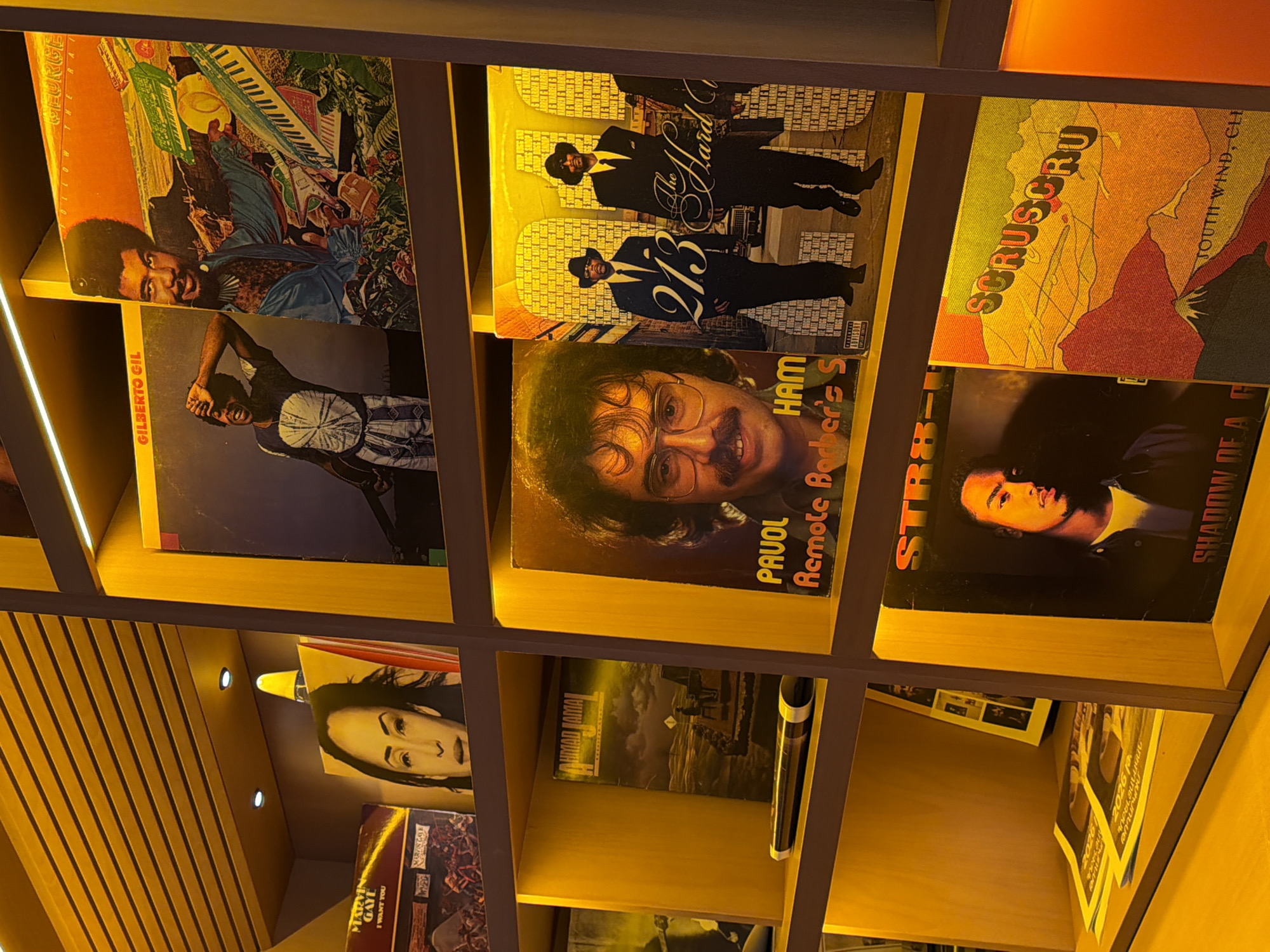 The image shows a shelf of vinyl records in a store. The store appears to be well-lit, with warm yellow lighting. The shelves are made of wood, and the records are arranged in rows. Some of the record covers are visible, including albums by Gilberto Gil, George Duke, Ahmad Jamal, and 213. There is also a record with the title "STR8" and another with "SCRUSCRU." The records are in good condition, and the store appears to be clean and organized. The overall impression is one of a well-stocked and inviting record store.
The image does not contain any specific details or clues that would indicate its location in Yerevan, Armenia.