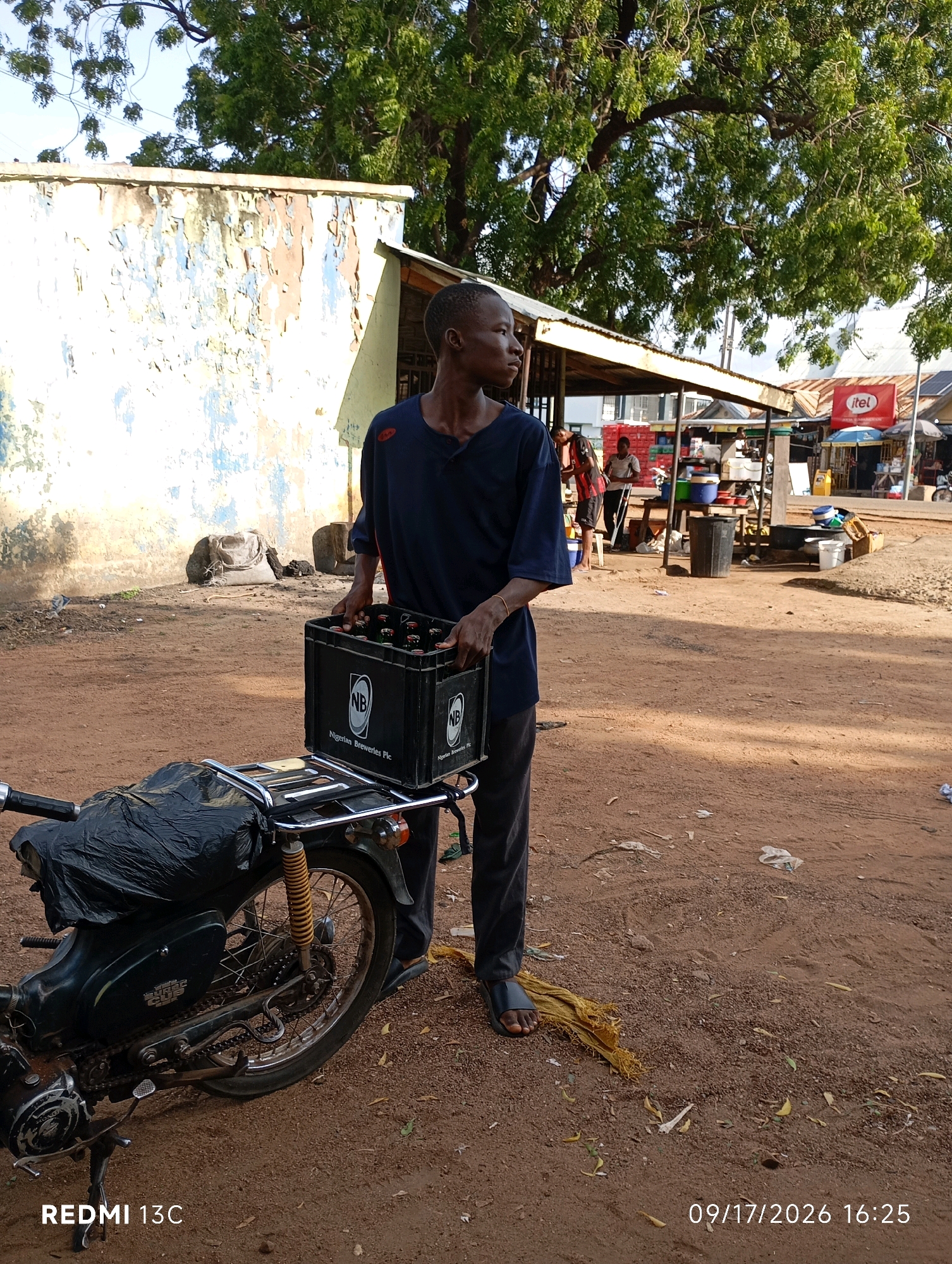 A young man in a dark blue shirt and grey pants stands in a dusty, outdoor environment, holding a black plastic crate filled with bottled beverages. The crate is marked with the "NB" logo and "Nigerian Breweries Plc". He is positioned next to a motorcycle, which has a black plastic bag draped over its rear seat. In the background, there is a weathered building with peeling paint on the left, and a market area with stalls, umbrellas, and other people on the right. The scene is illuminated by natural light, suggesting it is daytime. The ground is covered in reddish-brown dirt and scattered debris. A timestamp and the text "REDMI 13C" are visible in the bottom left corner of the image. The setting appears to be a marketplace or a busy street in the city of Shendam, Nigeria, given the visual cues and the presence of the Nigerian Breweries product.