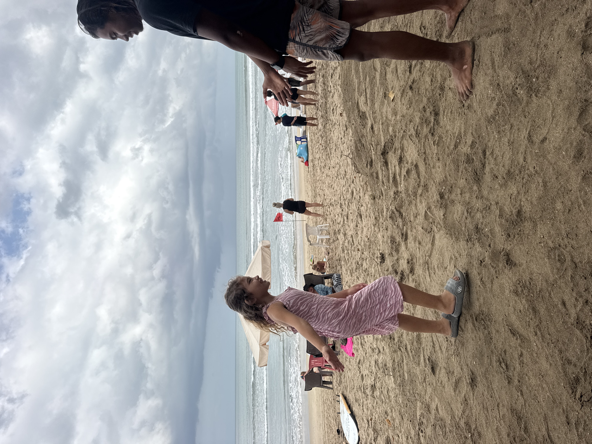In Kuta, Indonesia, on a cloudy day, a young girl with wavy brown hair and wearing a pink striped dress is standing on the beach, facing a man. The man, with dark hair and wearing a black t-shirt and patterned shorts, stands to her right. The girl has her left arm extended. In the background, several people are visible on the sandy beach, some sitting under umbrellas or chairs, while others stand near the ocean. Waves are rolling in, and a surfboard lies on the sand to the girl's left. Red flags are planted in the sand, and beach umbrellas are set up.