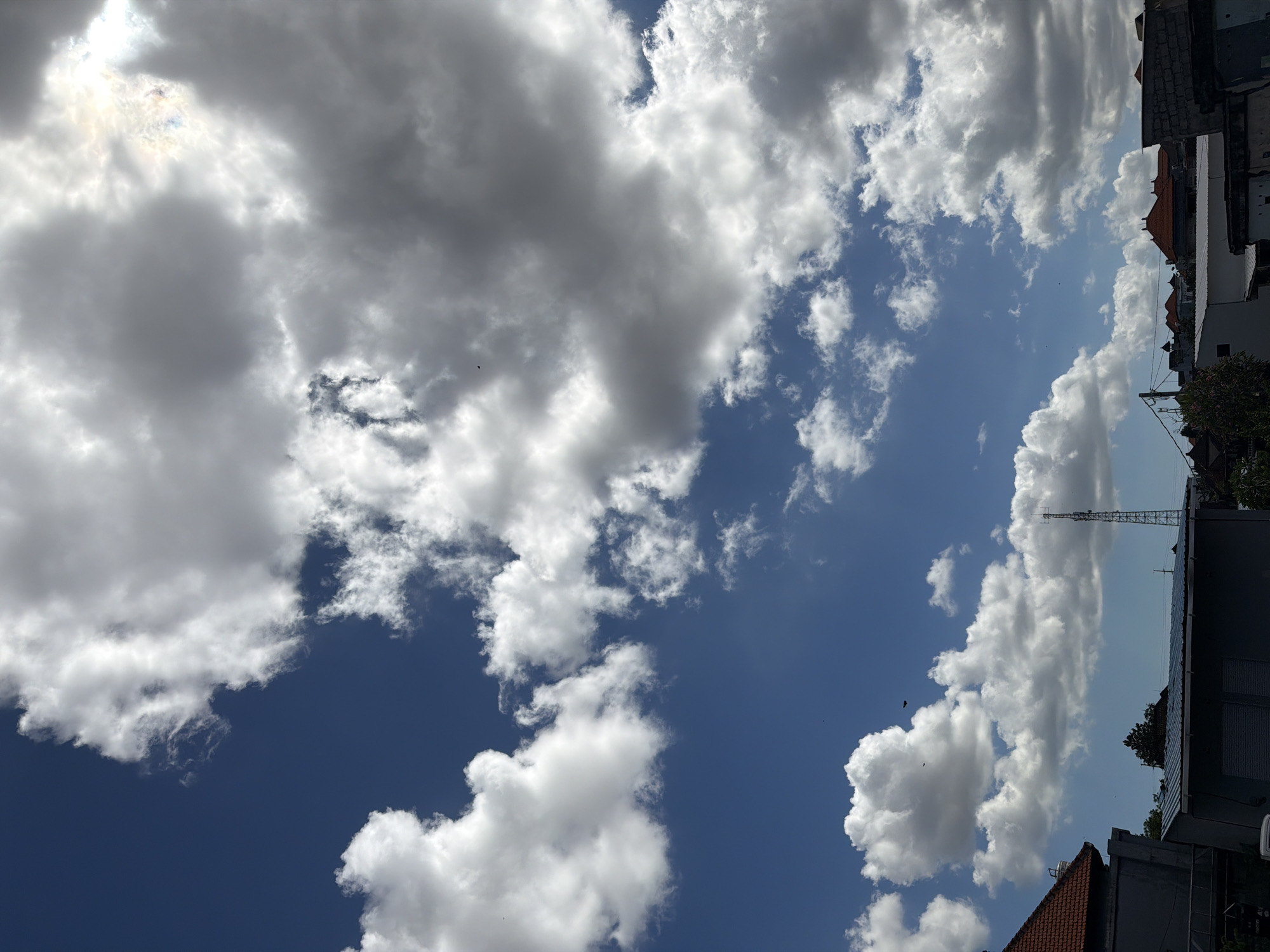 The image is an upward view of the sky with various types of clouds, including dark gray and fluffy white clouds, against a backdrop of blue sky, taken in Denpasar, Indonesia. Sunlight filters through a gap in the clouds on the top left. A communication tower is visible amidst the clouds on the right side of the image. The edges of rooftops and buildings are visible on the right side of the image. No people are visible. The sky appears to be active with cloud movement.
