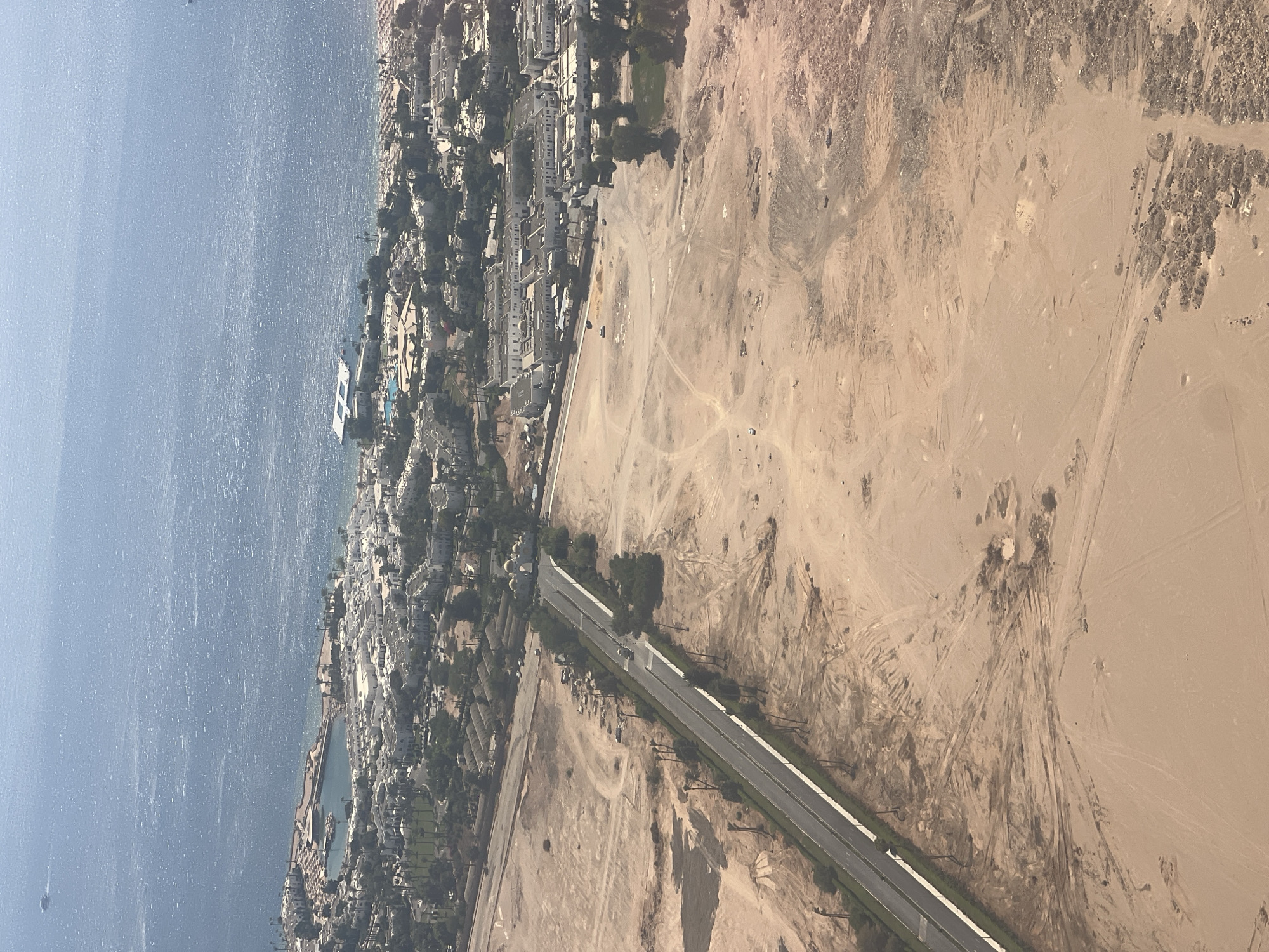 This aerial image shows a section of Sharm El Sheikh, Egypt, featuring a resort area along the coastline and undeveloped desert land inland. The sea is visible in the upper portion of the image, with a bright, rippling surface. Adjacent to the sea is a cluster of white resort buildings with red-tiled roofs, surrounding a bay or harbor with a pier. Further inland, the landscape transitions into sandy desert with sparse vegetation, including palm trees and some green areas within the resort. A paved road runs diagonally across the desert, bordered by a grassy median and palm trees. The desert terrain displays tire tracks and evidence of construction or land clearing. Several vehicles are present on the road and in the desert.