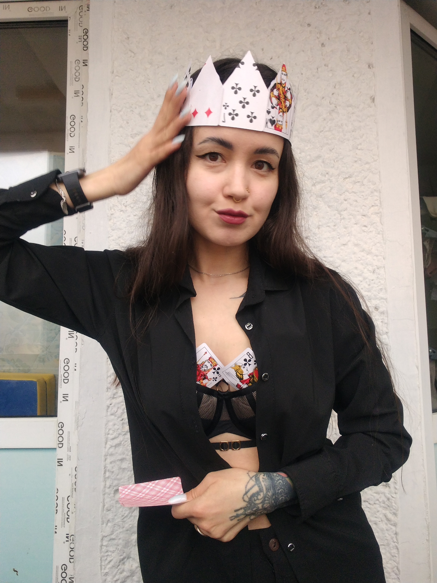 A young woman is captured in a half-body portrait, posing against a light-colored, textured wall next to a window. She has dark, long hair, dark eyebrows, winged eyeliner, and dark red lipstick, complemented by a gold nose ring. Her expression is a subtle smirk, looking directly at the viewer with confidence.
She wears a handmade paper crown crafted from playing cards, featuring suit symbols like spades, clubs, and diamonds, as well as face cards like the King of Hearts and Jack of Hearts. Her left hand is raised, lightly touching the crown. She is dressed in an unbuttoned black long-sleeved shirt or blazer, revealing a black bra underneath. The bra has a decorative element resembling a playing card pattern, with details of a King of Clubs visible. A silver chain necklace is also visible. On her left wrist, she wears a black digital watch and a silver bracelet. Her right forearm sports a dark, intricate tattoo. In her right hand, she holds a single playing card face down, showing a red patterned back.
The background features a white window frame with repeating text "GOOD IN" and the Cyrillic letter "И" (pronounced 'ee' or 'i') arranged vertically. Through the window, glimpses of blue and yellow objects are visible, suggesting an indoor or covered outdoor setting, possibly a balcony. The lighting is bright and natural, indicating daytime. The scene conveys a playful and assertive mood, with a strong playing card theme, potentially set in Noviy, Russia.