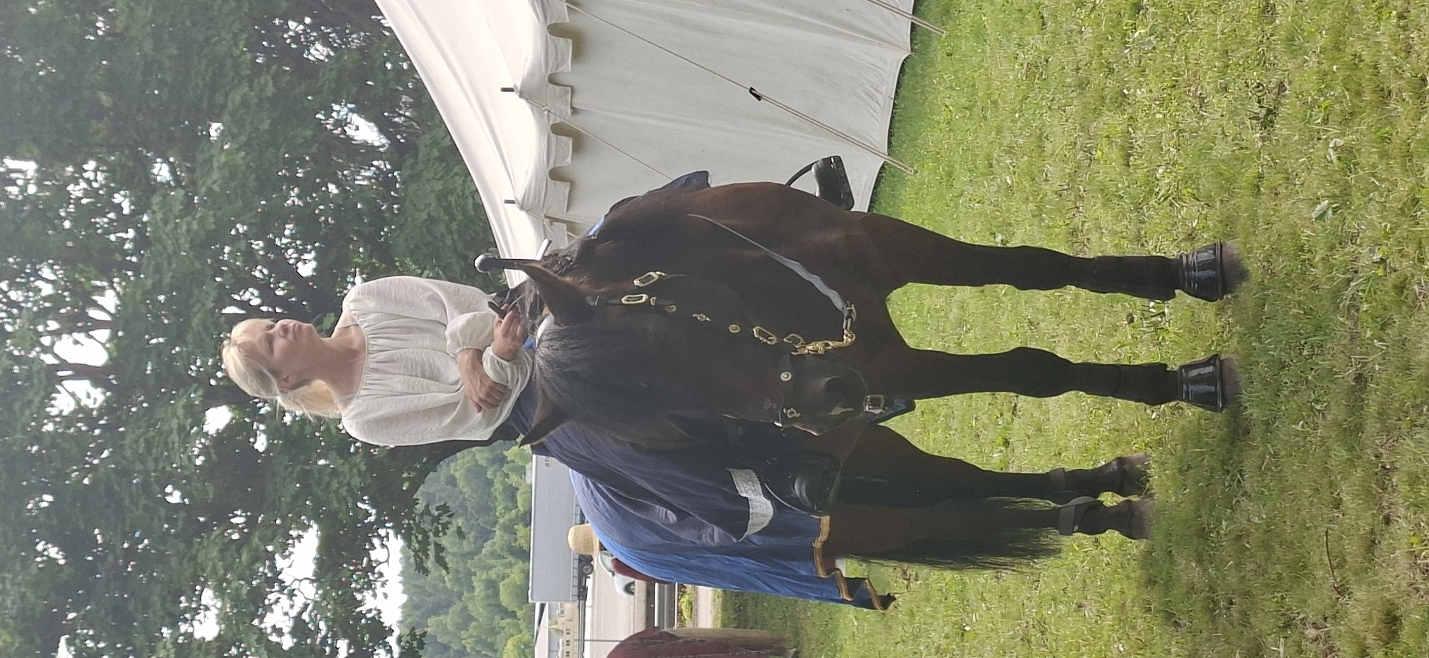 A woman with blonde hair pulled back in a ponytail, wearing a loose-fitting, off-white long-sleeved shirt, is seated on a dark brown horse. She is looking to her right. The horse is outfitted with a bridle and saddle, and a dark blue blanket with a white trim is draped over its back and sides. The horse is standing in a grassy field, with a large, light-colored tent visible in the background to the right. Behind the tent, a distant treeline is visible. In the far background, a highway and a large truck can be seen, suggesting the presence of a nearby road or transportation hub. The scene appears to be outdoors, possibly at a historical reenactment or festival, given the attire of the woman and the horse's tack. The weather appears to be overcast or hazy. The specific location in Turku, Finland cannot be determined from the visual information provided.