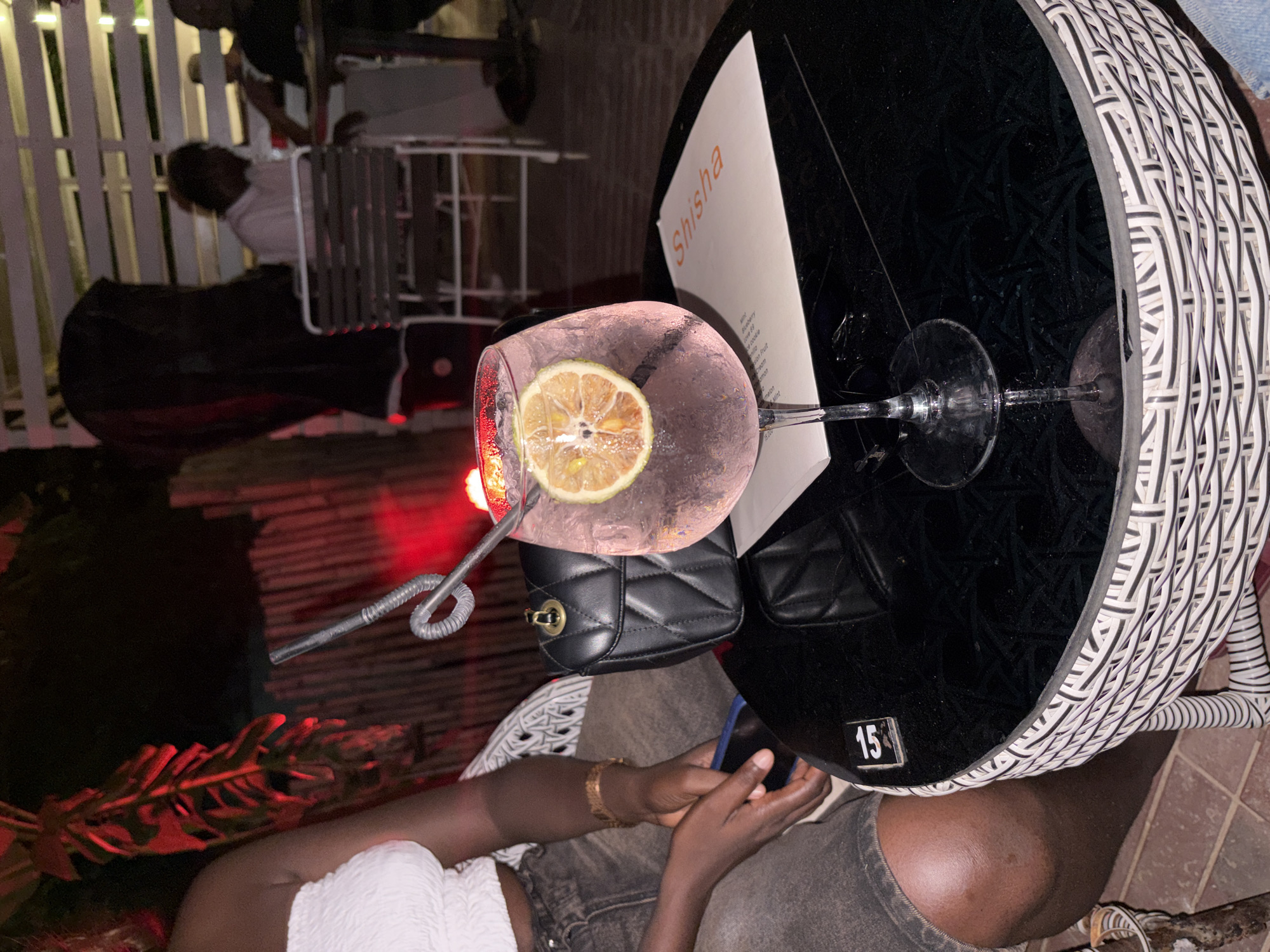 The image depicts a cocktail on a table in an outdoor setting, likely in Abuja, Nigeria, based on the "Shisha" menu visible. The cocktail is in a large glass with a lemon slice and a flexible straw. A small black purse is placed next to the cocktail. A person wearing a white top and jeans is sitting at the table, their hands visible, and appears to be holding a phone. The table is black and surrounded by a woven wicker-style chair. A number "15" is affixed to the side of the table. In the background, there is a white picket fence, a person sitting on a white chair, and ambient red lighting near plants. The overall scene appears to be at a bar or lounge.
