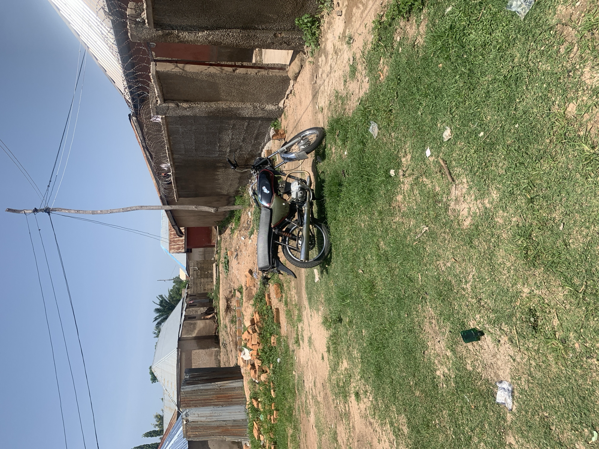 The image depicts an outdoor scene in Jauro Yinu, Nigeria. A black and olive green motorcycle is parked on a dirt path surrounded by dry grass and some patches of green grass. The motorcycle has a passenger seat and a metal rack above the rear wheel. In the background, several residential structures are visible. One building has a corrugated metal roof and concrete walls. Another structure features a grey concrete wall topped with barbed wire, and a red door or entrance is partially visible. To the left, there are piles of red bricks and a corrugated metal sheet structure. The ground is a mixture of soil and sparse vegetation. A wooden pole with electrical wires stretches across the top of the frame. The sky is clear and blue.