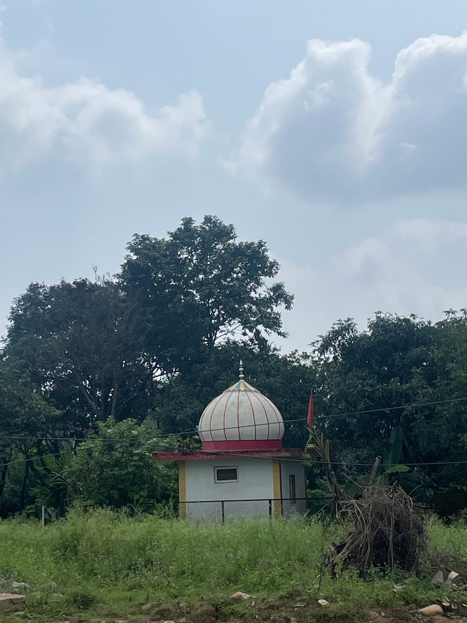 The image shows a small, white temple with a red and white domed roof, located in Shahpur, India. The temple is surrounded by lush green vegetation and tall trees. The sky is overcast with large, white clouds. A red flag is visible on top of the temple. In the foreground, there is a pile of dry branches and roots. The overall scene depicts a serene and tranquil environment.