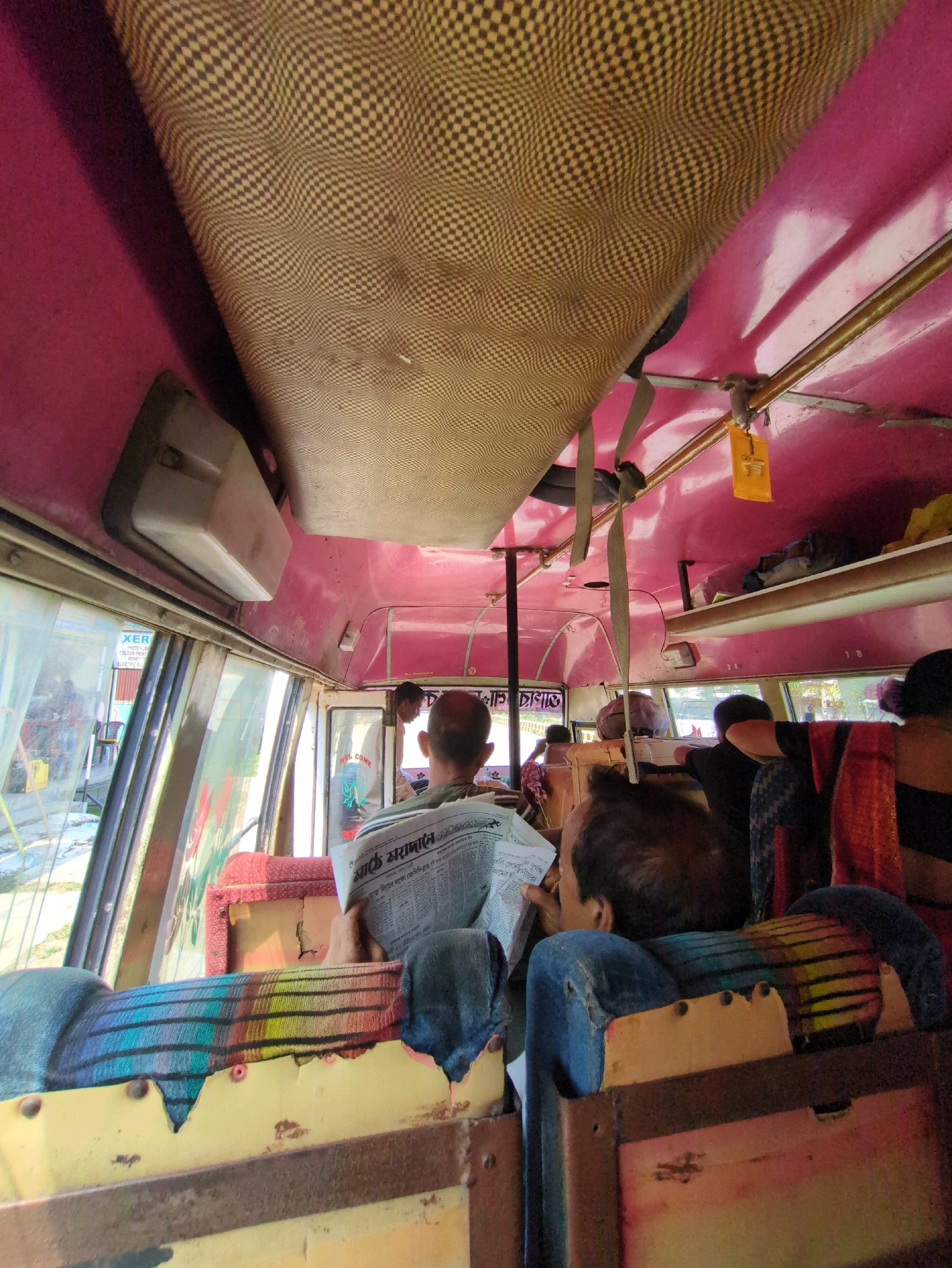 The image shows the interior of a bus. The ceiling is covered with a tan checkered fabric. The walls and ceiling are painted pink. There are overhead shelves with various items. A man is seated in the foreground, reading a newspaper. Another person's head is visible to the right, with a colorful cloth draped over their shoulder. The seats appear to be covered with striped fabric. There are windows on the side of the bus showing an outdoor scene. A white electrical fixture is mounted on the ceiling near the front. A yellow tag with some text is hanging from a metal rod. The overall impression is of a rustic and worn interior of a public transportation vehicle. The scene is located in Kumarghat, India.
