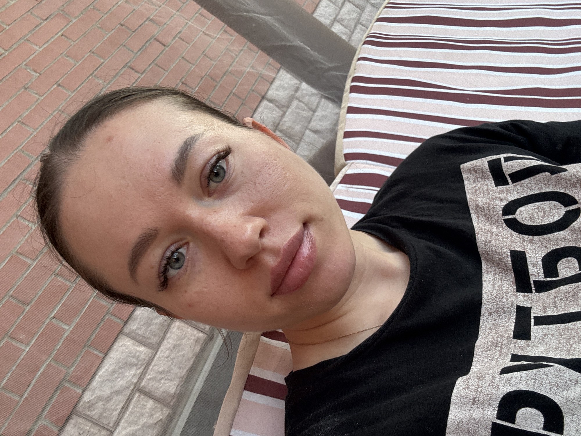 The image is a selfie of a young woman with fair skin, light-colored eyes, and dark hair pulled back. She is wearing a black t-shirt with white lettering and is lying on a striped surface, possibly a mattress or lounge chair. Behind her to the left, there is a brick wall and a gray paved area. The location is possibly in or near Ферма, Russia, based on the prompt, though there is no verifiable evidence of the exact location within the image itself. The woman's gaze is directed towards the camera, and her expression is neutral.