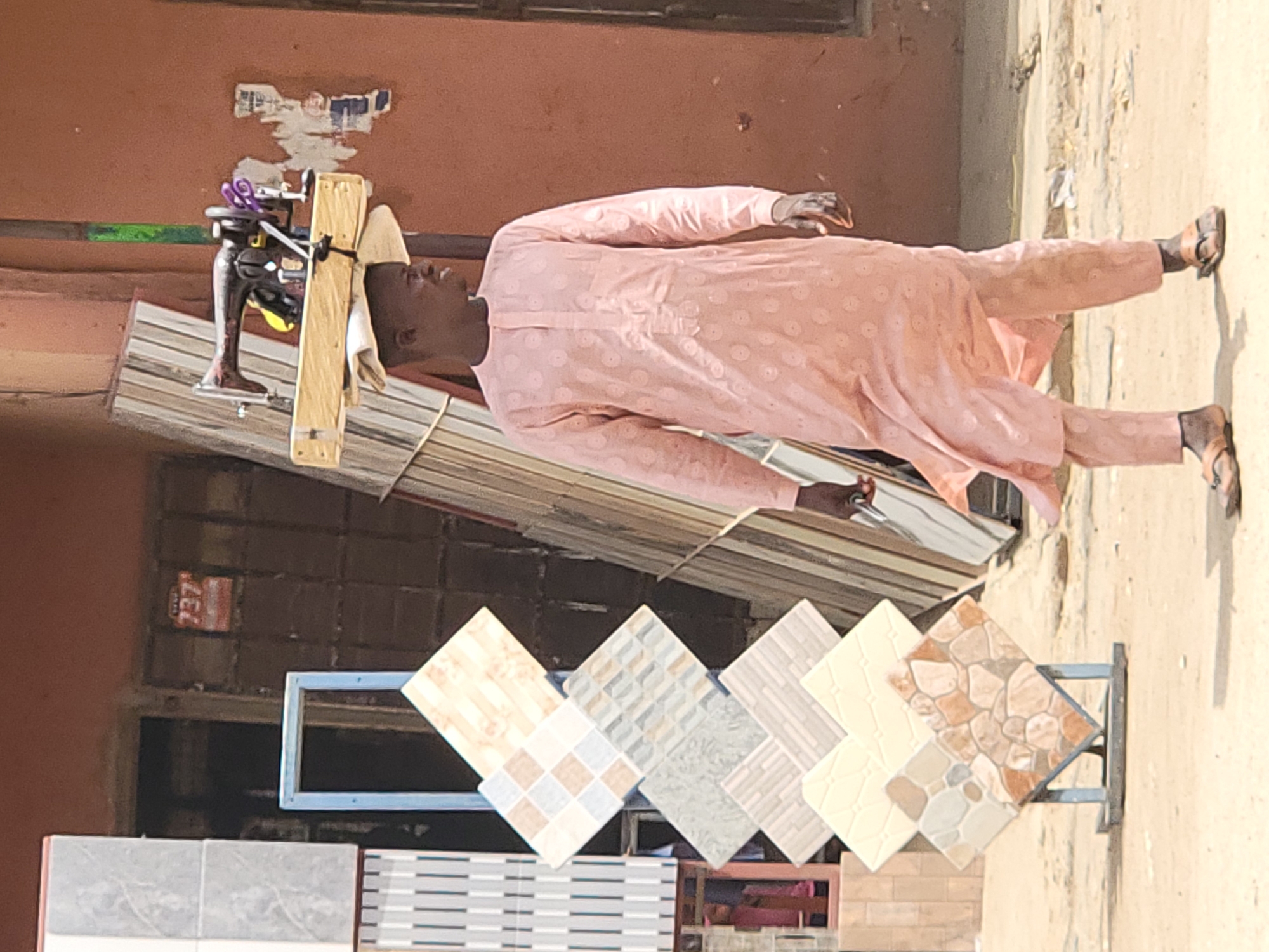 The image, taken in Maiburiji, Nigeria, captures an outdoor street or market scene on a bright, sunny day. The primary subject is a dark-skinned man walking on an unpaved, sandy path. He is dressed in a light peach-colored, long-sleeved tunic (likely a traditional Nigerian agbada) and matching trousers, both featuring a subtle embroidered circular pattern. He wears simple brown sandals.
The man is notably carrying a wooden platform on his head, a common method of transport in the region. Attached to this platform is an old, dark metal hand-cranked machine, possibly a sewing machine or a similar mechanical device, with some purple fabric or rope resting on it. This suggests he may be a street vendor or craftsman moving his equipment.
In the foreground, several display stands showcase various ceramic tiles. These square tiles exhibit diverse patterns, including geometric designs, textured surfaces, and natural stone-like finishes, indicating a tile seller's display in a commercial area.
The background features a reddish-brown wall, likely part of a building. On this wall, torn pieces of paper or posters are visible, and a small red rectangular sign with the white numbers "237" is clearly discernible. Above the man, a corrugated metal roof or awning is partially visible. The scene conveys a glimpse into daily life and commerce in the area.