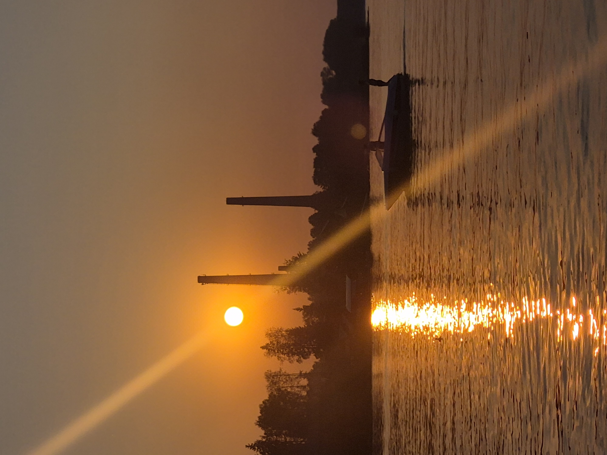 The image captures a sunset over the Kama River in Chaikovsky, Russia. Two industrial chimneys, silhouetted against the orange sky, stand on the far bank, accompanied by a line of trees. The sun, a bright orb, is low in the sky, casting a brilliant reflection on the water's surface. A small motorboat with two visible figures is navigating the river, heading towards the right side of the frame. The overall scene depicts a tranquil, atmospheric evening on the river.