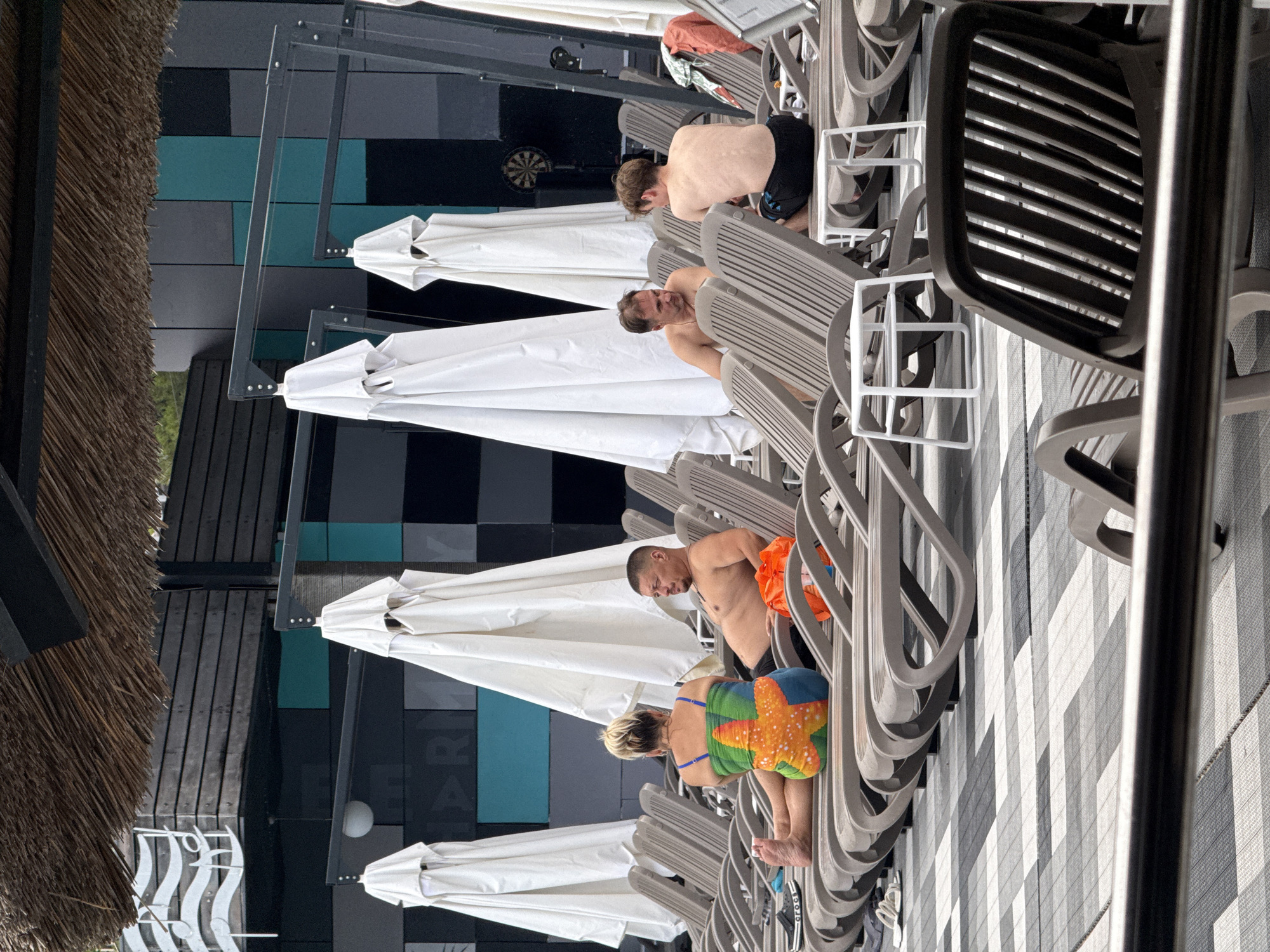 A group of people are relaxing on lounge chairs by a pool. The scene appears to be at a resort or hotel. A woman in a colorful swimsuit is sitting on a lounge chair, facing away from the camera. A man with a shaved head is sitting in a lounge chair next to her, looking towards the right. Another man is sitting on a lounge chair further to the right, also facing away from the camera. In the background, there are several white umbrellas, a building with a dark facade and geometric patterns, and a dartboard. The ground is covered with a patterned tile. The overall atmosphere is one of leisure and relaxation.