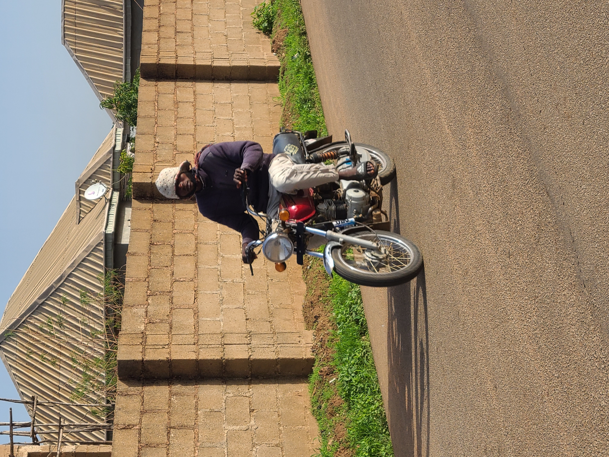 A man is riding a red motorcycle on a paved road next to a tall brick wall in an urban setting. The man is wearing a dark long-sleeved shirt, light-colored pants, sandals, and a white knit cap. The road surface is coarse and there is green vegetation along the side of the road and at the base of the wall. The brick wall has visible seams between the bricks. Beyond the wall, a building with a corrugated metal roof is visible under a light blue sky. The city and specific location are unidentified.