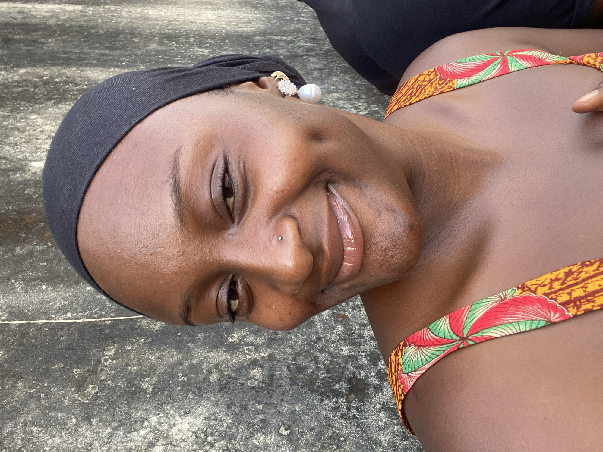 A woman is lying on a concrete surface, possibly the ground, in New Stadium, Nigeria. She is wearing a black headwrap, earrings, and a garment with yellow, red, and green patterned straps. She is smiling and facing the camera. Another person's legs are visible in the background. The concrete surface is slightly weathered and textured.