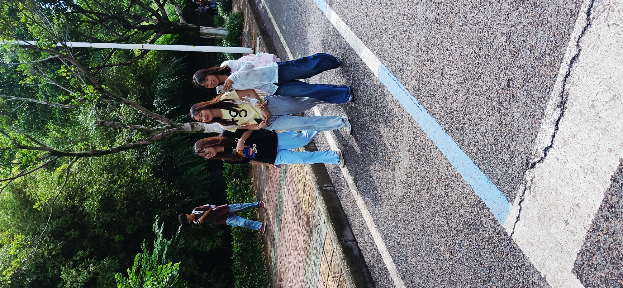 The image shows a group of three young women walking in single file along a road or paved area. The woman at the front is wearing a light pink or white button-down shirt with blue jeans. The woman in the middle is wearing a yellow shirt with the number "8" on it and gray pants. The woman at the back is wearing a black shirt and light blue jeans. They appear to be chatting and possibly posing for a photo.
The location appears to be outdoors, adjacent to a lush green area filled with trees and dense foliage. To the side of the women is a paved walkway made of reddish-brown and yellow tiles. Next to the walkway is a road with a blue painted line.
Based on the metadata, the image was taken in Chongqing High-tech Zone, China. A person walking along the brick path can be seen behind the young women.