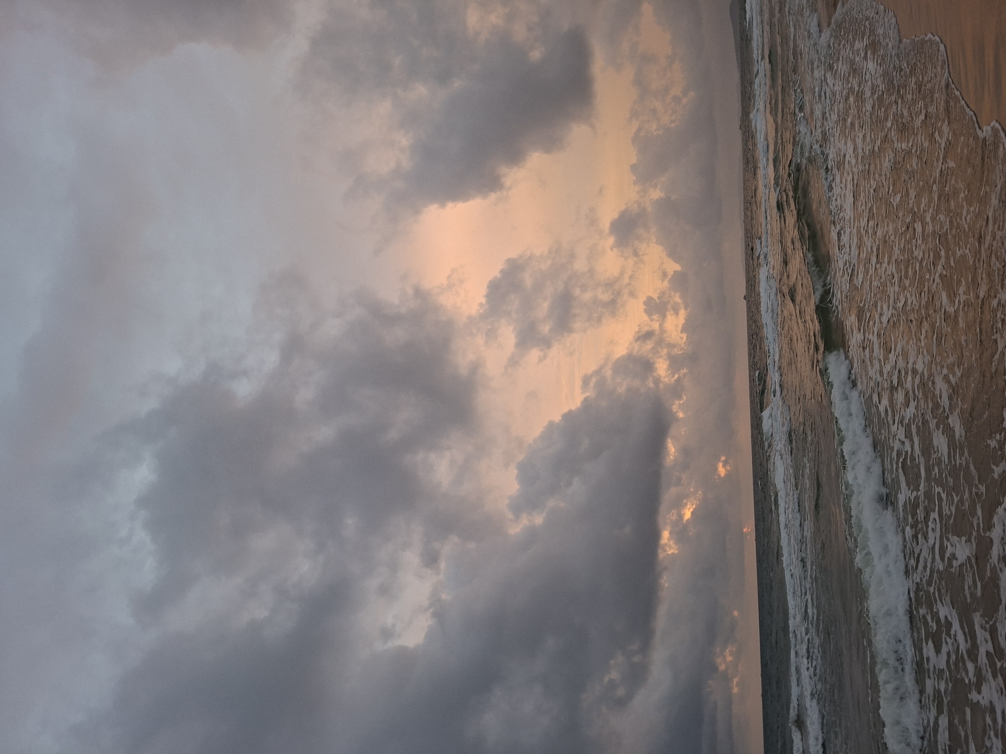The image depicts a beach scene in Phú Quốc, Vietnam, at dusk. The sky is filled with heavy, gray clouds that are tinged with a soft, peachy orange light, suggesting the sun is setting or has recently set. The sea is choppy with white-capped waves rolling towards a sandy shore. The water reflects the muted colors of the sky, creating a somber yet beautiful atmosphere. No people are visible in the image, and the scene is dominated by the natural elements of sky and sea. The overall impression is one of a dramatic and atmospheric coastal landscape.