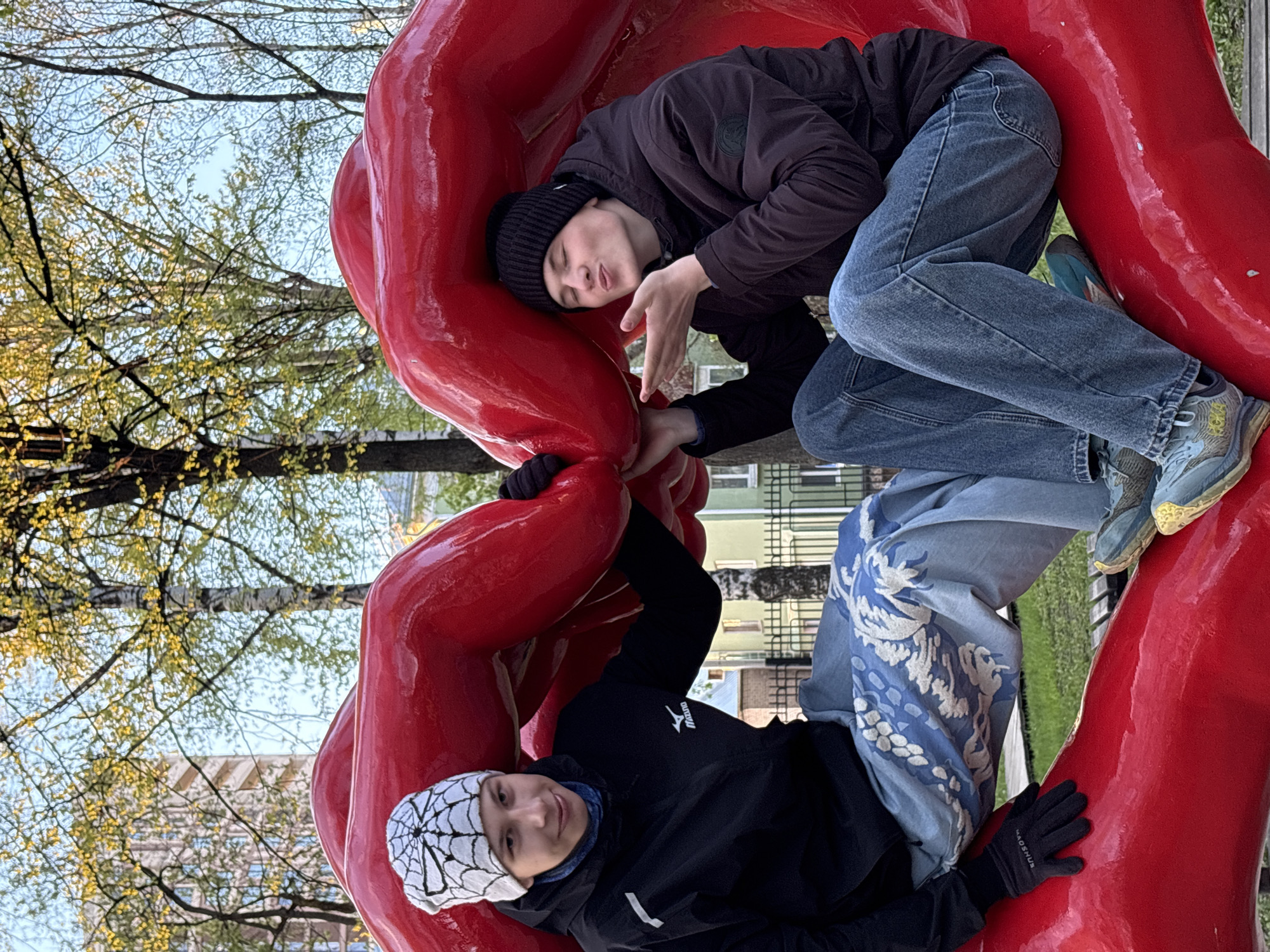 Two people are reclining within the open, bright red sculpture of a pair of lips located in Perm, Russia. The sculpture is positioned outdoors, surrounded by trees and buildings. The person on the left wears a black jacket, gloves, and a spiderweb-patterned beanie. The person on the right wears a dark jacket, blue jeans, and a dark beanie.