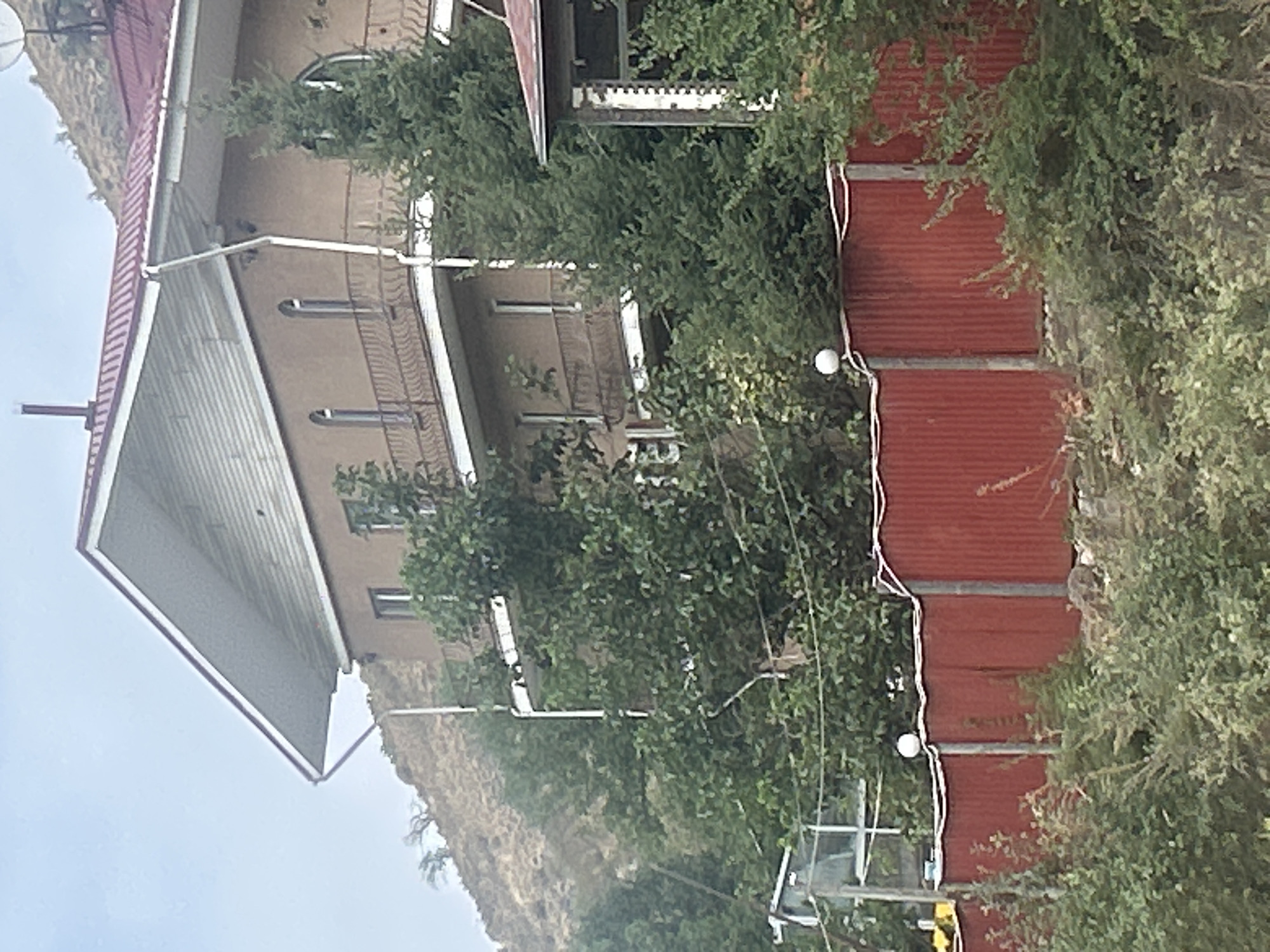The image depicts a multi-story building situated on a hillside. The building has a brown stucco exterior and a red metal roof. There are balconies with ornate railings on at least two levels. The windows are arched on the upper floors. Evergreen trees and dense foliage surround the building, obscuring parts of its lower levels. A red corrugated metal fence runs across the foreground, with white decorative edging and vertical posts. A white spherical light fixture is visible on top of the fence. In the background, a barren, dry hillside rises steeply. The sky is overcast and pale. The location is identified as Koklaisai, Kazakhstan.