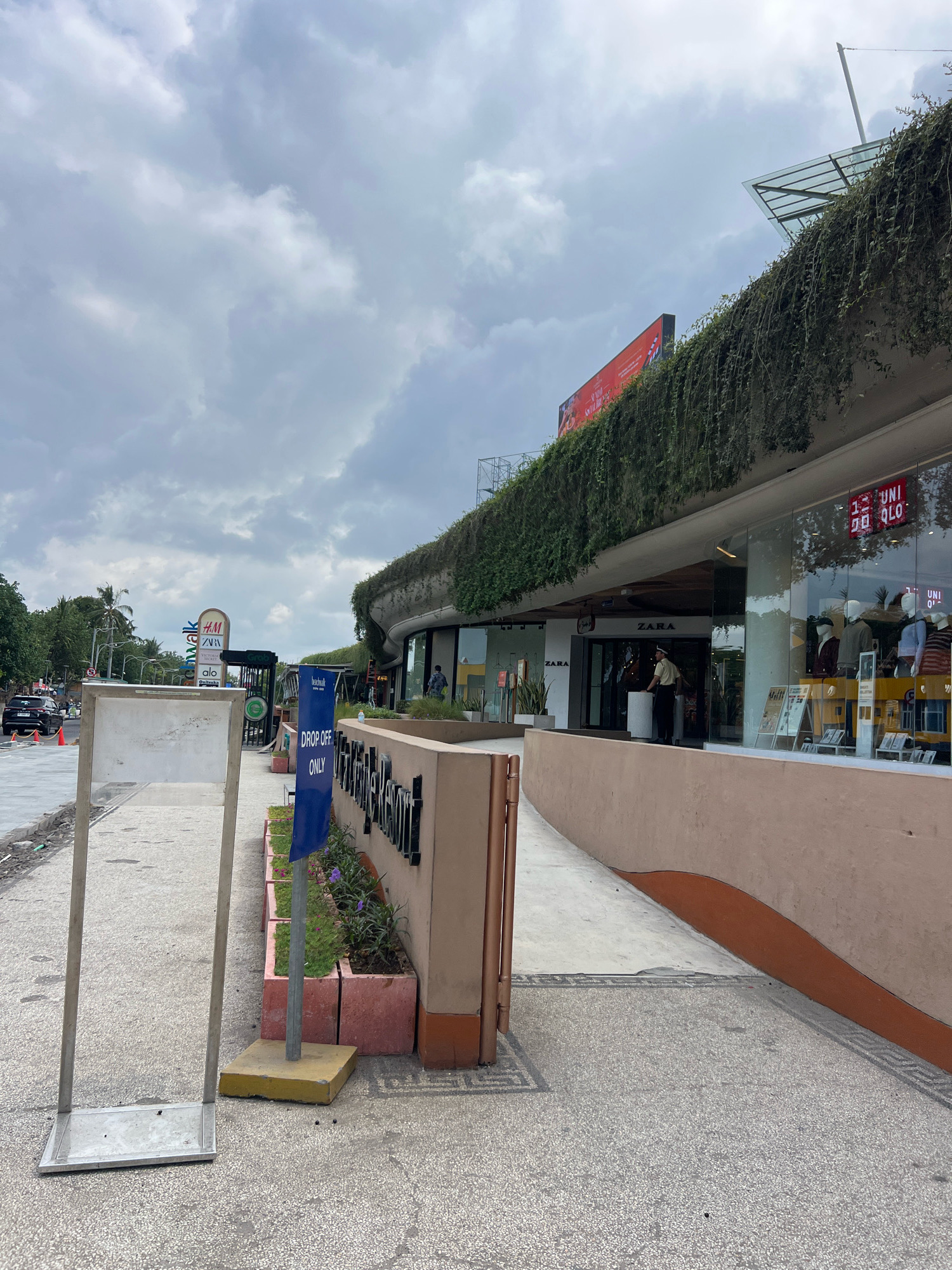 The image shows the exterior of a modern shopping complex in Kuta, Indonesia, under a cloudy sky. The building features a curved facade covered in lush green vegetation, creating a unique architectural style. In the foreground, there is a sidewalk with a textured surface. A silver metal sign stand is positioned on the left side of the sidewalk, and a blue "DROP OFF ONLY" sign is visible on a pole further down.
Several storefronts are visible, including "ZARA" and "UNIQLO," indicated by their prominent signage and window displays featuring mannequins dressed in clothing. A person in a uniform, possibly a security guard or employee, stands near the entrance of the ZARA store.
To the left of the shopping complex, a street is visible with a few cars and trees lining the background. A signpost displays logos for several brands, including H&M, ZARA, and others. The overall scene suggests a bustling commercial area in Kuta.