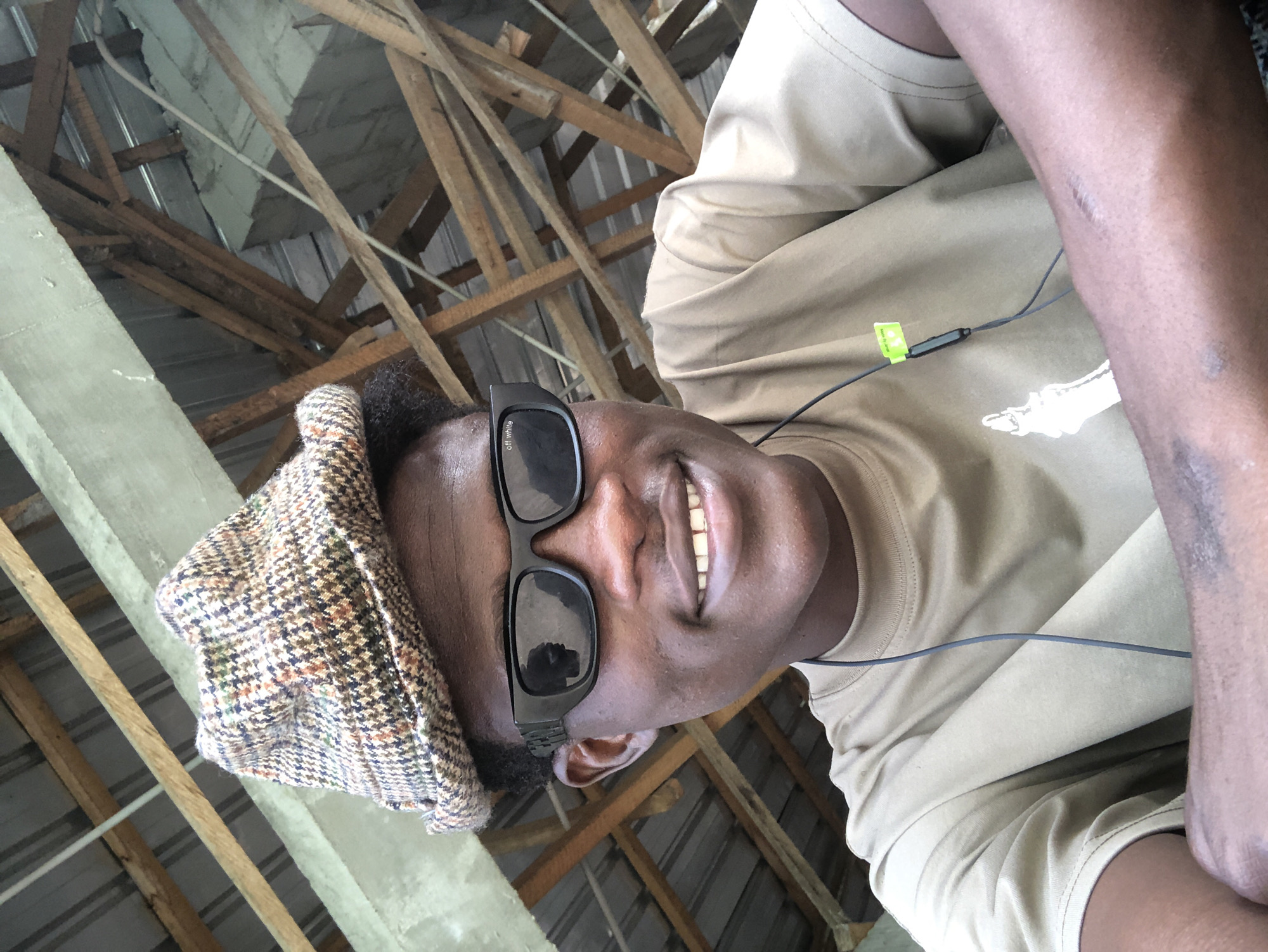 In Ado, Nigeria, a young Black male is captured in a close-up, waist-up selfie taken from a low angle. He is wearing a brown and tan plaid bucket hat, black sunglasses with the "off white" logo visible, and a plain tan t-shirt. He has a visible scar on his left forearm. The background consists of a wooden roof structure. He is smiling and looking directly at the camera, with his arms resting in front of him. A pair of black earbuds with a lime green clip are attached to his shirt.