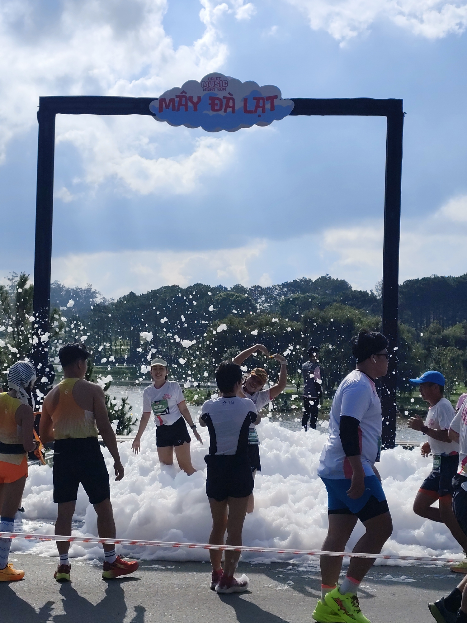 The image displays an outdoor daytime scene of a foam running event in Da Lat, Vietnam, identified as a "Dalat Music Night Run." A prominent, dark-colored rectangular archway frames the central activity, featuring a cloud-shaped sign on its top crossbar that reads "DALAT MUSIC NIGHT RUN" (in smaller text) and "MÂY ĐÀ LẠT" (Cloud Da Lat) in larger red lettering. Below the archway, a substantial volume of white, bubbly foam covers the ground, reaching above the knees of many participants. Numerous individuals, mostly adults, are present within and around the foam zone.
Participants are dressed in athletic attire, including t-shirts, shorts, and running shoes. Some are actively interacting with the foam; two women in the center, smiling, are partially submerged in the foam, with one forming a heart shape with her hands. Other individuals are seen walking or standing, observing the scene. On the far left, a person wearing orange shorts, a yellow top, and a striped head covering stands with their back to the camera. On the far right, a man in a white t-shirt and blue cap is visible in profile. Foam bubbles are suspended in the air, indicating active foam generation.
In the mid-ground, behind the foam, a body of water (likely a lake or large pond) is visible, reflecting the bright sky. Beyond the water, a dense line of trees forms a forested backdrop, with further hills and vegetation in the distance. The sky above is light blue with scattered white cumulus clouds. The ground beneath the foam appears to be a paved surface, with an orange and white striped barrier tape or rope visible in the immediate foreground.