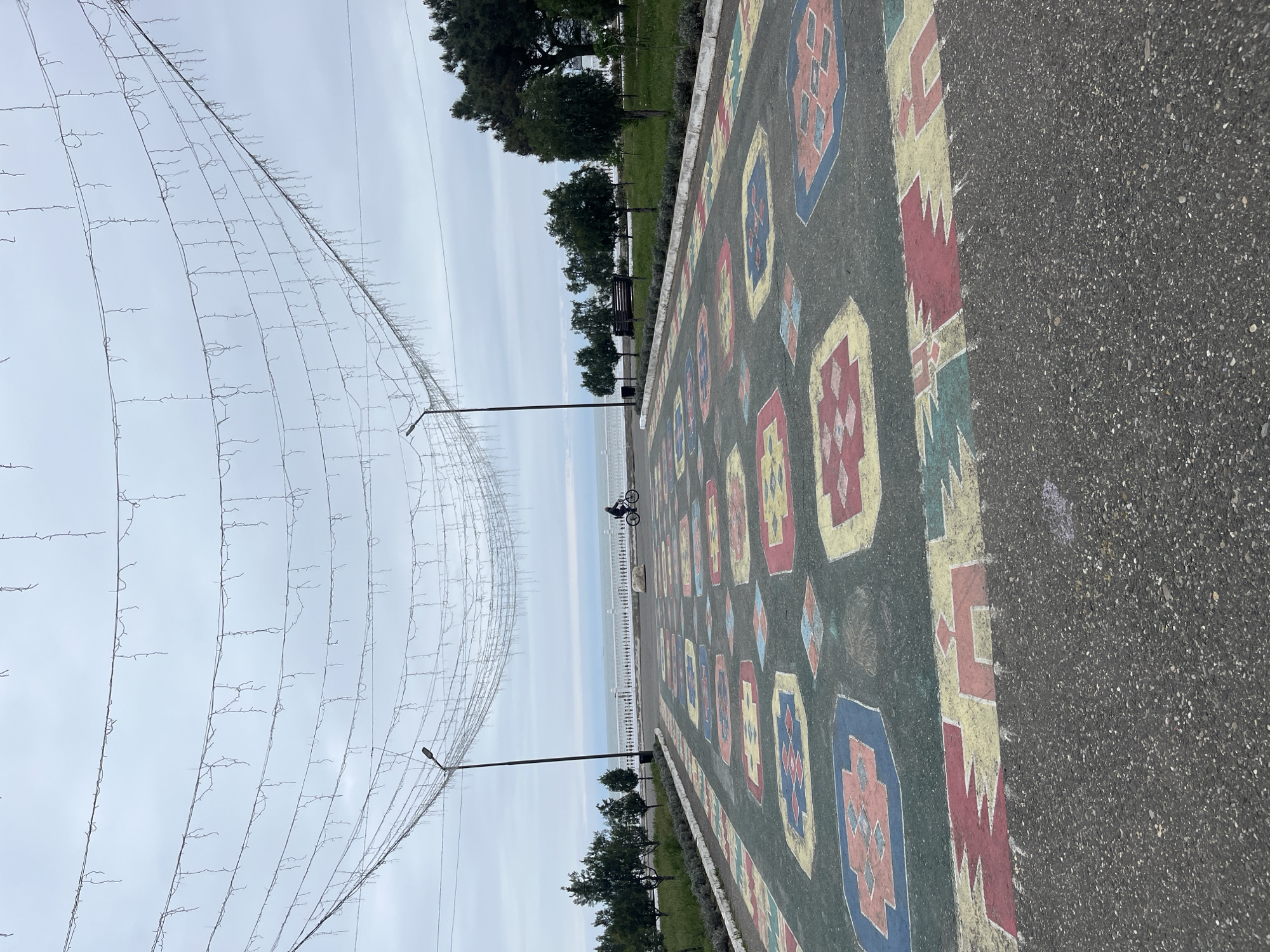 The image shows a section of a pedestrian walkway in Kaspiysk, Russia. The walkway is decorated with a painted pattern resembling a traditional carpet. A person on a bicycle is riding along the walkway in the distance. To the left, strings of bare light bulbs are suspended above the walkway. On the other side of the walkway, there is a strip of greenery with trees and benches facing towards what appears to be a body of water. The sky is overcast.