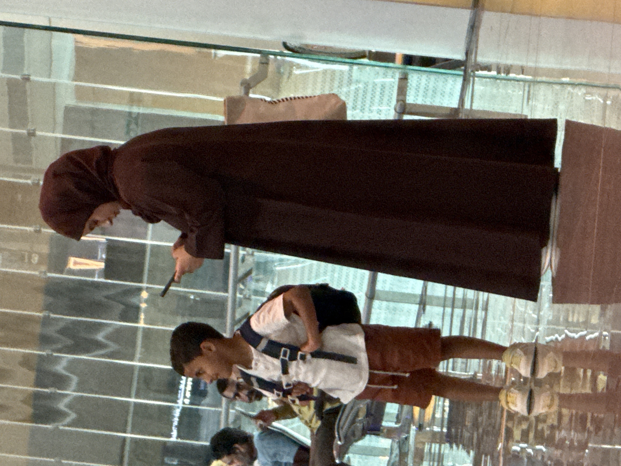 A young boy in a white t-shirt, orange shorts, and a backpack stands on a reflective floor. He is looking to his left, and his right hand is on his backpack. To his right, a woman wearing a dark headscarf and a dark robe is looking down at her phone. In the background, there are several people sitting and standing in an indoor area with large windows. The scene appears to be a public space, possibly an airport terminal, in Seeb Province, Oman.