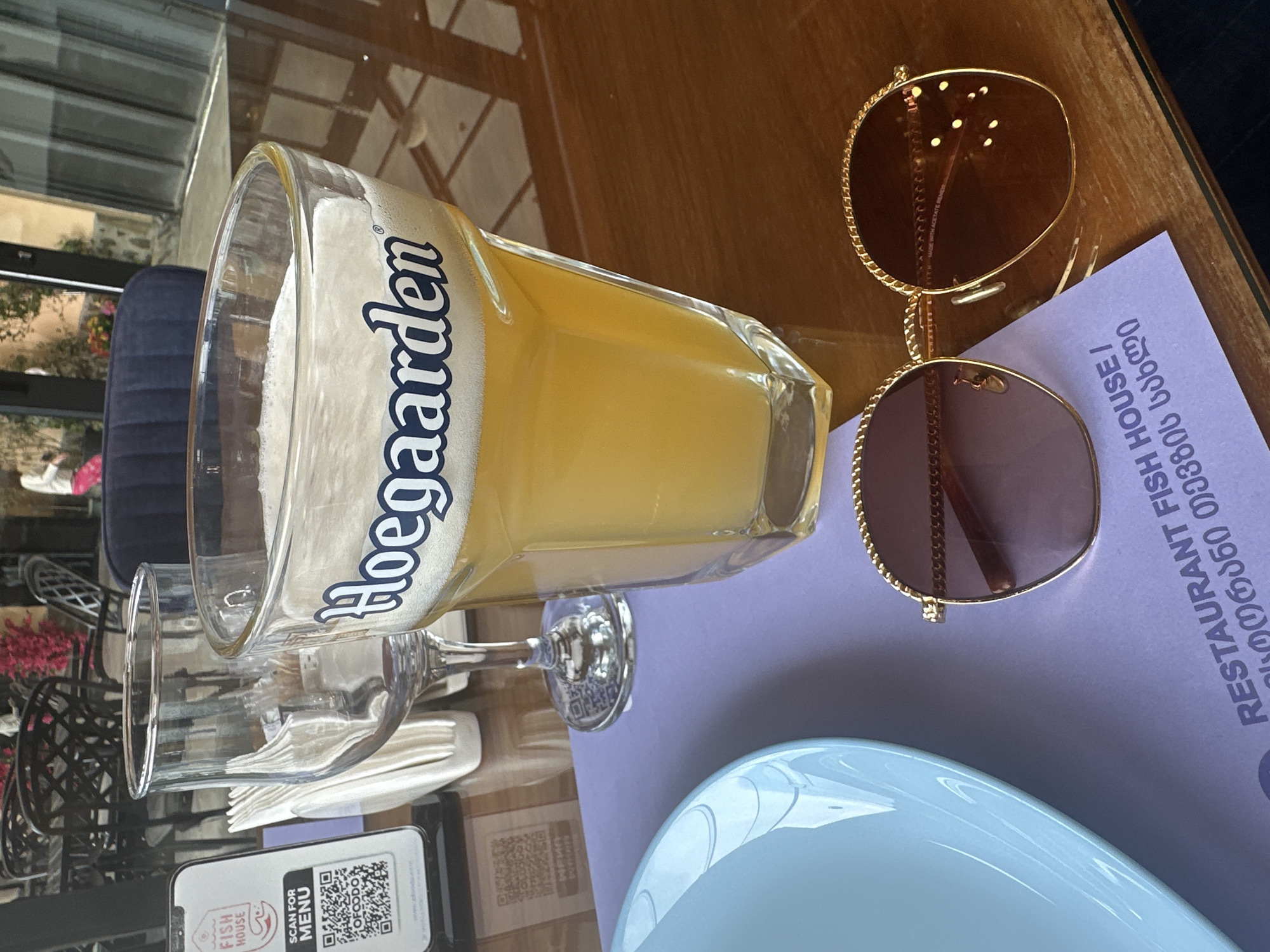 The image depicts a table setting with a glass of Hoegaarden beer, a pair of sunglasses, a blue plate, a phone with a QR code, and a restaurant menu. The menu identifies the restaurant as "RESTAURANT FISH HOUSE" and includes text in Georgian. The location of the restaurant within the city of Rekha, Georgia cannot be confirmed from the image.