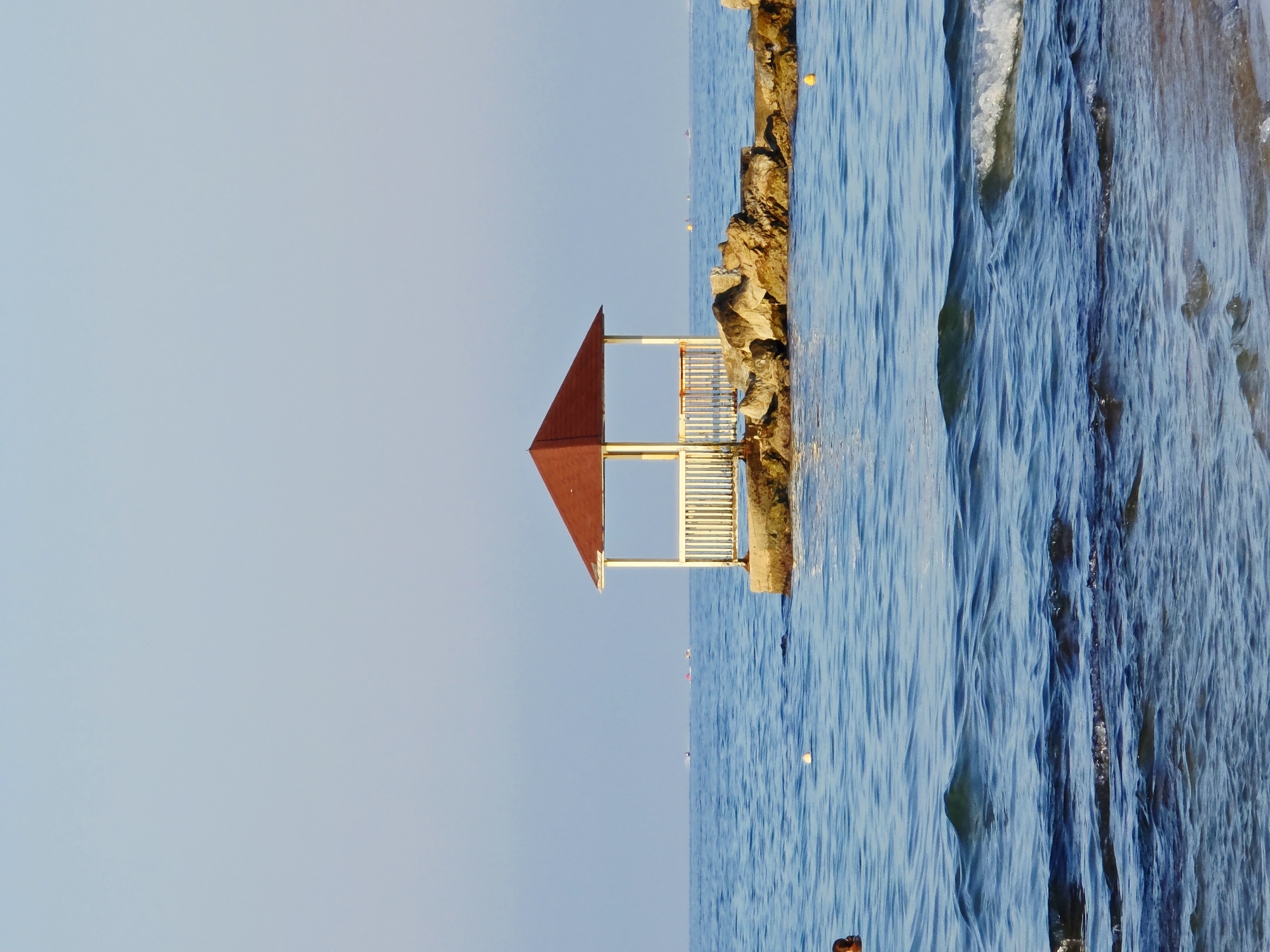 A gazebo with a red roof and white railings sits on a rocky outcropping in the clear blue sea. The water is calm with small waves breaking near the shore in the foreground. Several boats are visible in the distance. A person is partially visible swimming in the water to the left of the image. The sky is clear and pale blue. The scene appears to be a tranquil beach setting in Malia Municipal Unit, Greece.